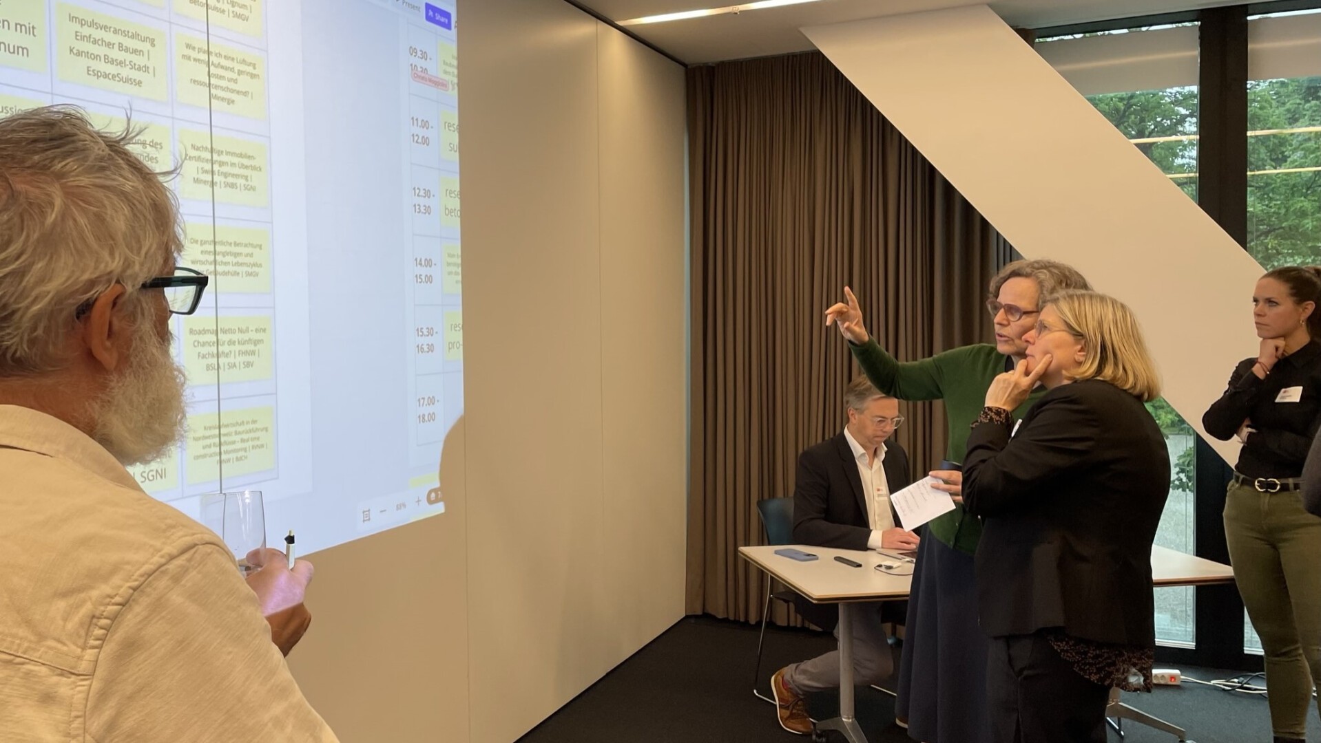 Vier Personen befinden sich in einem Besprechungsraum. Drei Personen stehen und diskutieren über Themen, die auf einer Projektionsfläche angezeigt werden, während eine Person an einem Tisch sitzt und sich Notizen macht. Die Atmosphäre wirkt konzentriert und kooperativ.