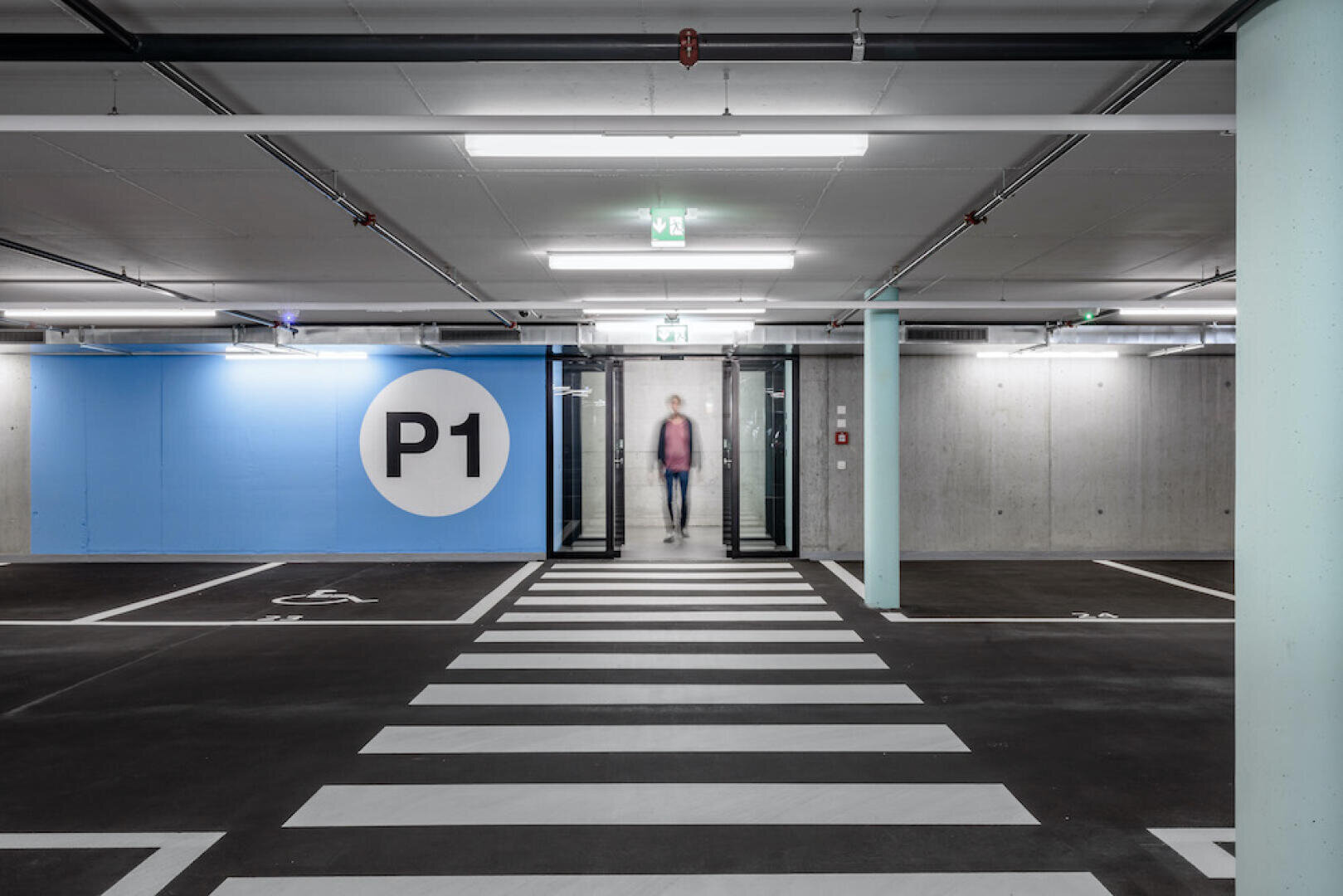 Eine moderne Tiefgarage mit einer blauen Wand mit der Aufschrift "P1", weißen Parklinien, einem Fußgängerüberweg und einer Person, die durch Glastüren im Hintergrund auf die Kamera zugeht.