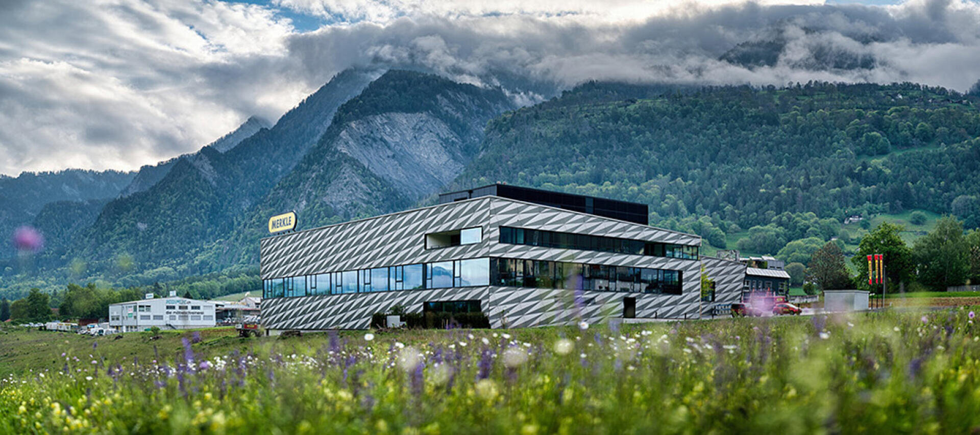 Ein modernes Gebäude aus Glas und Metall mit geometrischen Mustern steht in einem Grasfeld mit Wildblumen, hinter dem sich grüne, bewaldete Berge unter einem dramatischen Wolkenhimmel befinden.