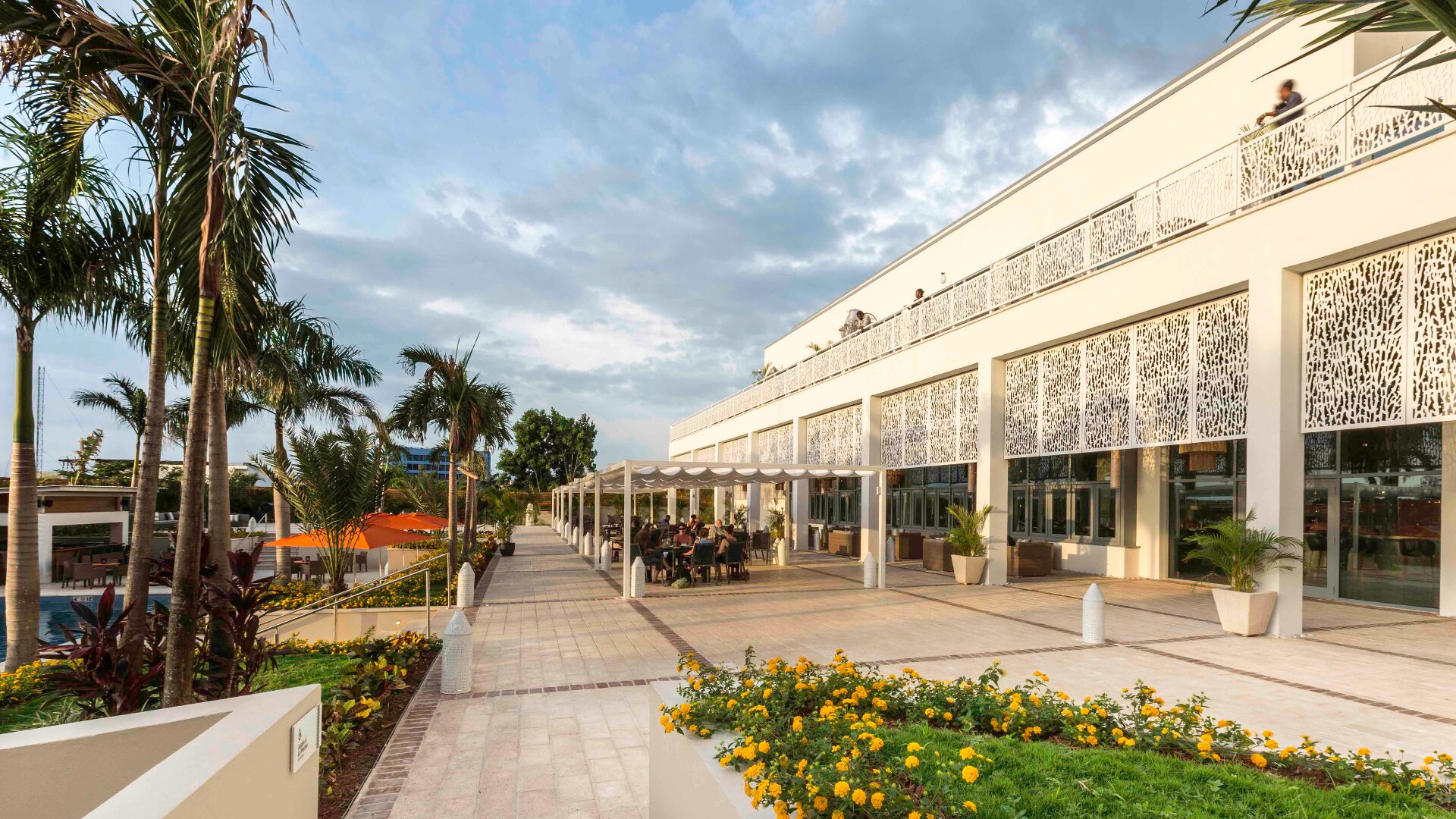 Ein modernes Gebäude mit dekorativen weißen Paneelen und großen Fenstern ist von Palmen und gelben Blumen umgeben. Die Menschen sitzen an Tischen auf einer großzügigen Terrasse unter einer Pergola, der Himmel ist bewölkt.