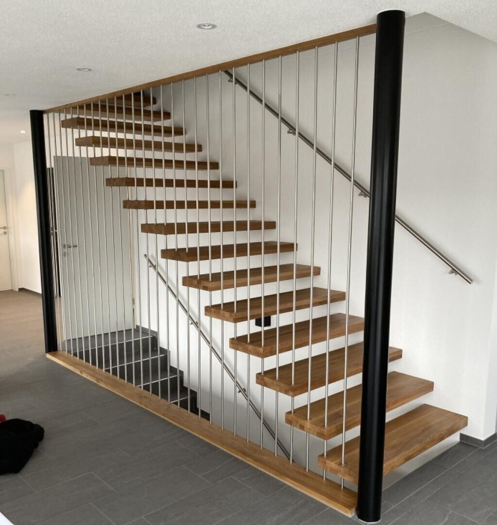 Escalier flottant moderne en bois avec des barreaux métalliques et un cadre vertical noir, devant un mur blanc avec un sol carrelé gris. L'escalier n'a pas de contremarches, ce qui donne une impression d'ouverture et d'aération.