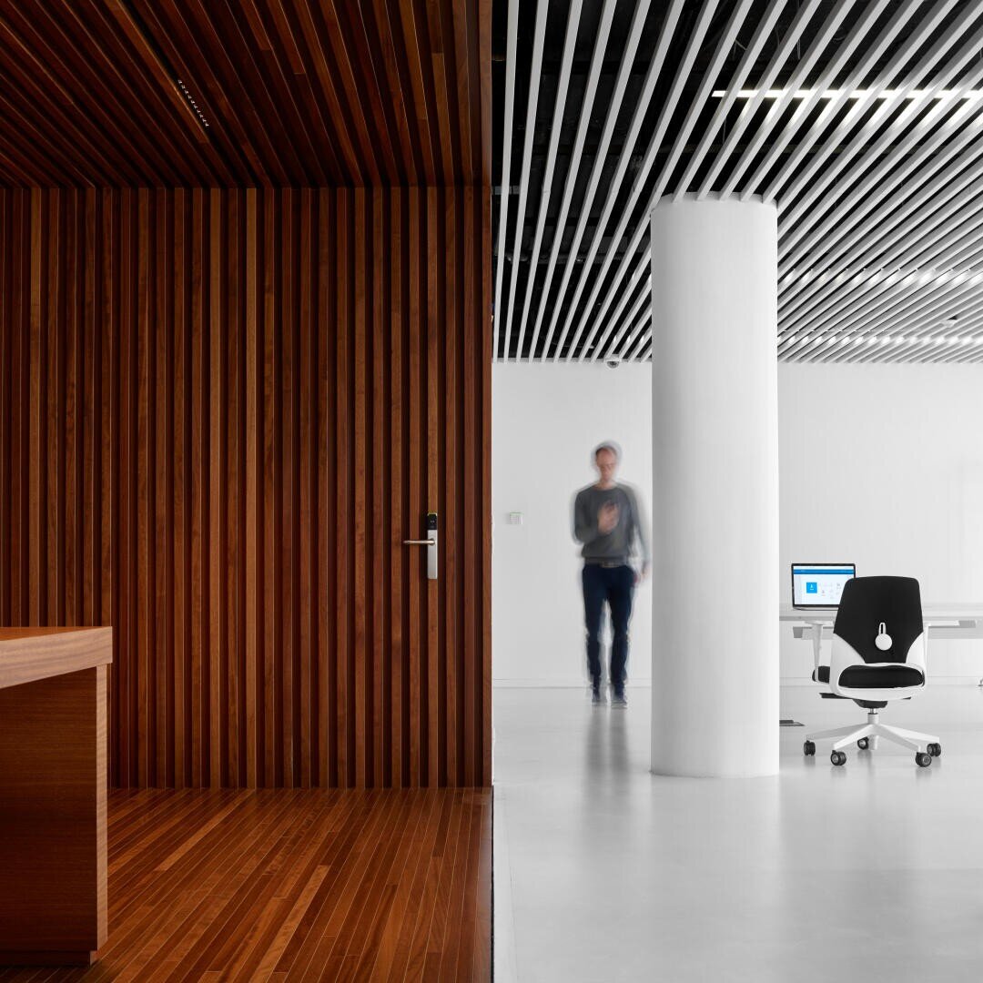 Un espace de bureau moderne, divisé par un mur en bois sur la gauche et une colonne blanche sur la droite, avec une personne floue en train de marcher, un bureau blanc et un ordinateur dans un environnement lumineux et minimaliste.
