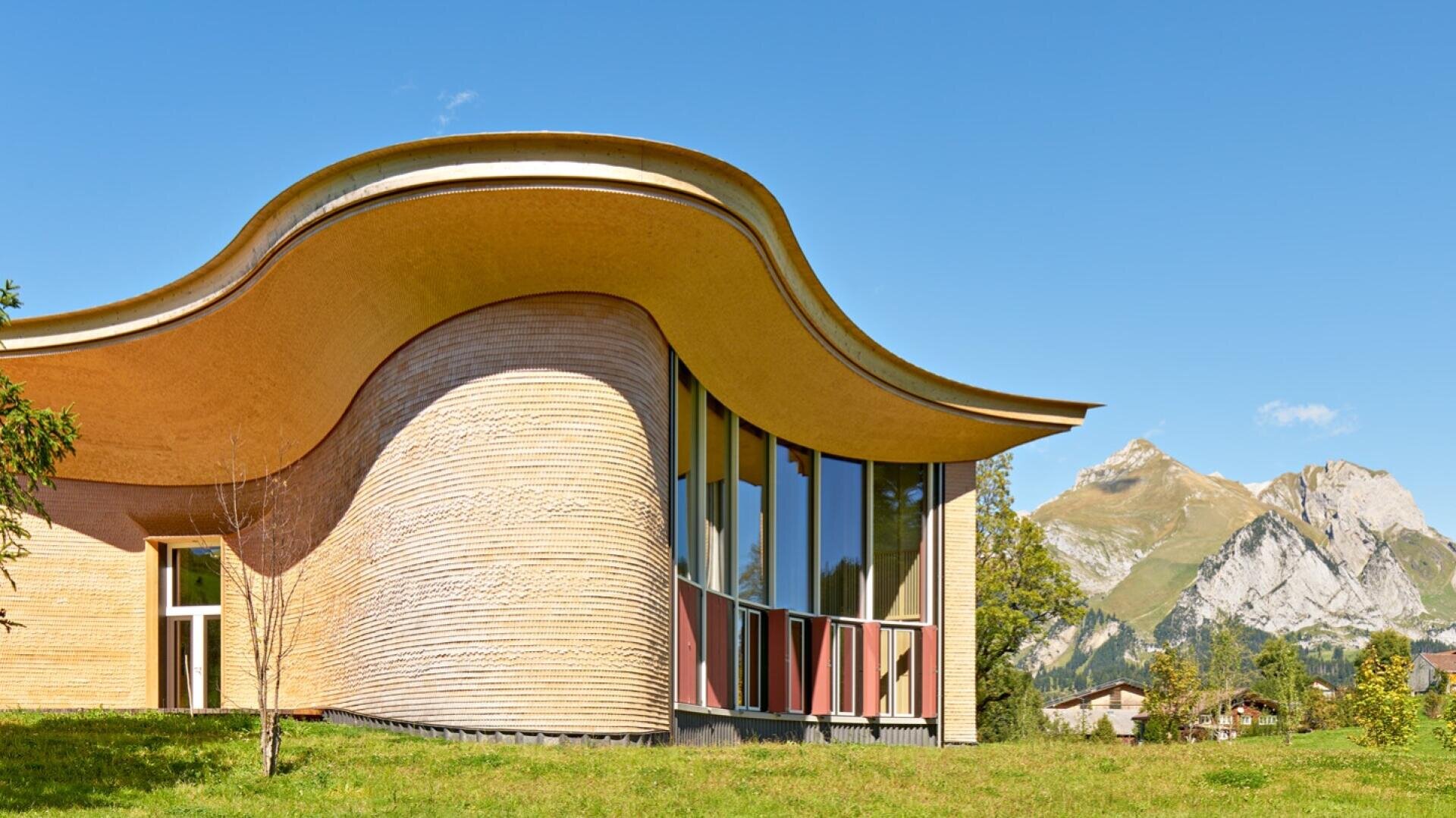 Ein modernes Gebäude mit einem gewellten, überhängenden Holzdach und großen Fenstern steht auf einer grasbewachsenen Wiese, mit Bergen und einem klaren blauen Himmel im Hintergrund.