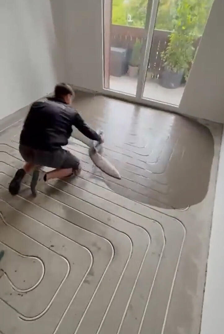 In einem hellen Raum mit großen Fenstern verteilt eine Person Spachtelmasse auf einem Boden mit freiliegenden Fußbodenheizungsrohren.