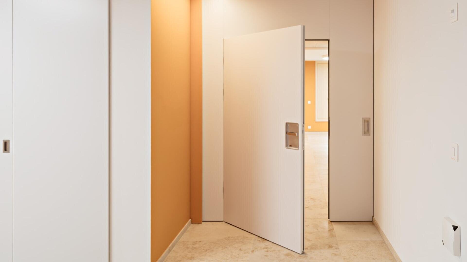 Ein moderner Innenflur mit beigen Böden, weißen Wänden, einer Schiebetür auf der linken Seite und einer weißen Flügeltür, die teilweise geöffnet ist und einen Raum mit warmen gelb-orangenen Wänden freigibt.