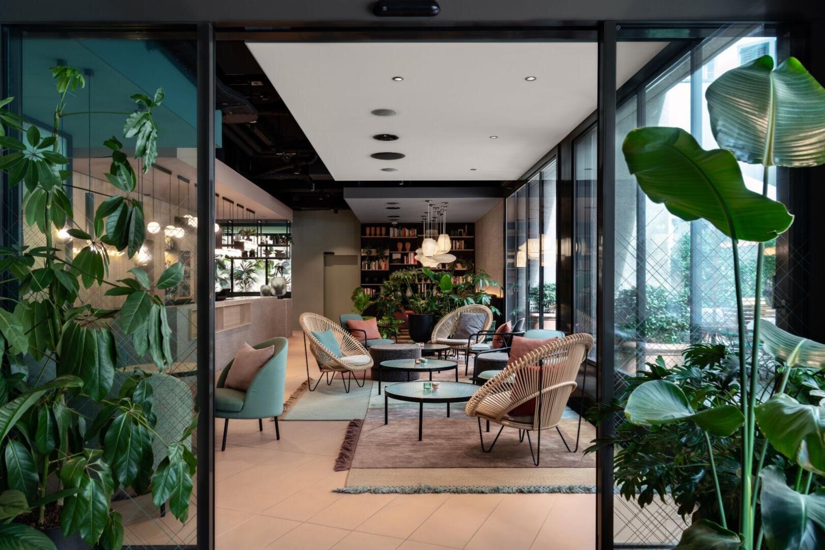 Salon moderne avec des chaises en rotin, des tables rondes, des coussins aux couleurs pastel et des plantes vertes luxuriantes. De grandes fenêtres laissent entrer la lumière naturelle et des étagères avec des livres et des lampes décoratives créent une atmosphère confortable et accueillante.