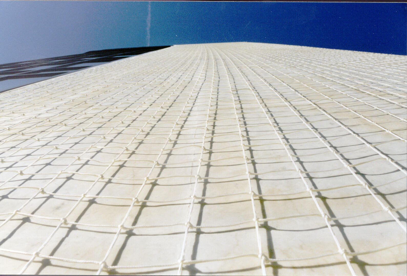Blick aus geringer Höhe auf eine hohe Struktur, die mit einem weißen Netz mit Gittermuster bedeckt ist, vor einem klaren blauen Himmel. Die Perspektive unterstreicht die Höhe und die vertikalen Linien der Struktur.