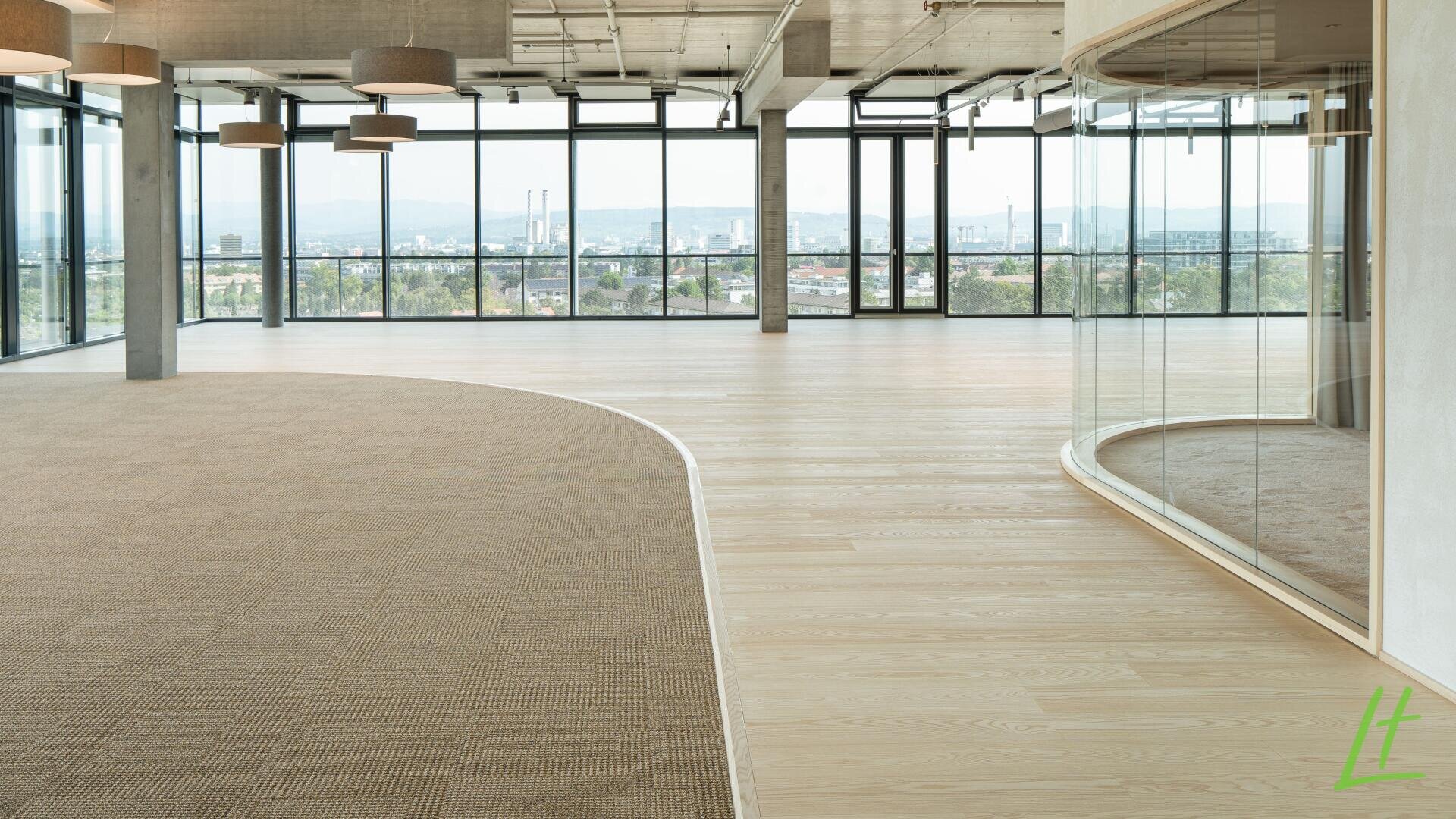 Moderner, leerer Büroraum mit großen Fenstern, Blick auf die Stadt, Holz- und Teppichböden, Deckenleuchten und einer gebogenen Glaswand auf der rechten Seite. Natürliches Licht durchflutet den offenen, geräumigen Raum.