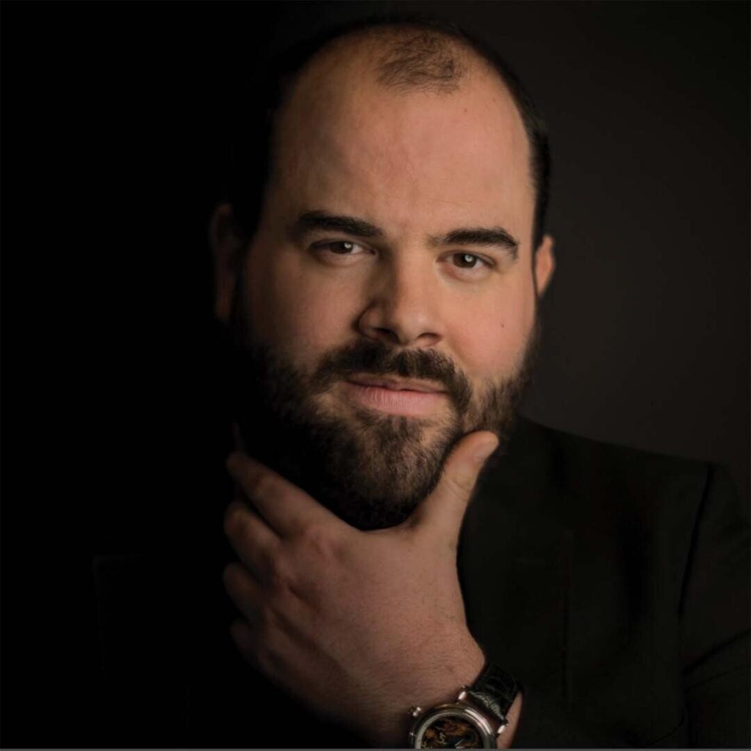 Un homme barbu à la chevelure dégarnie regarde la caméra en posant son menton sur sa main. Il porte une veste sombre et une montre, avec une expression neutre sur un fond sombre.