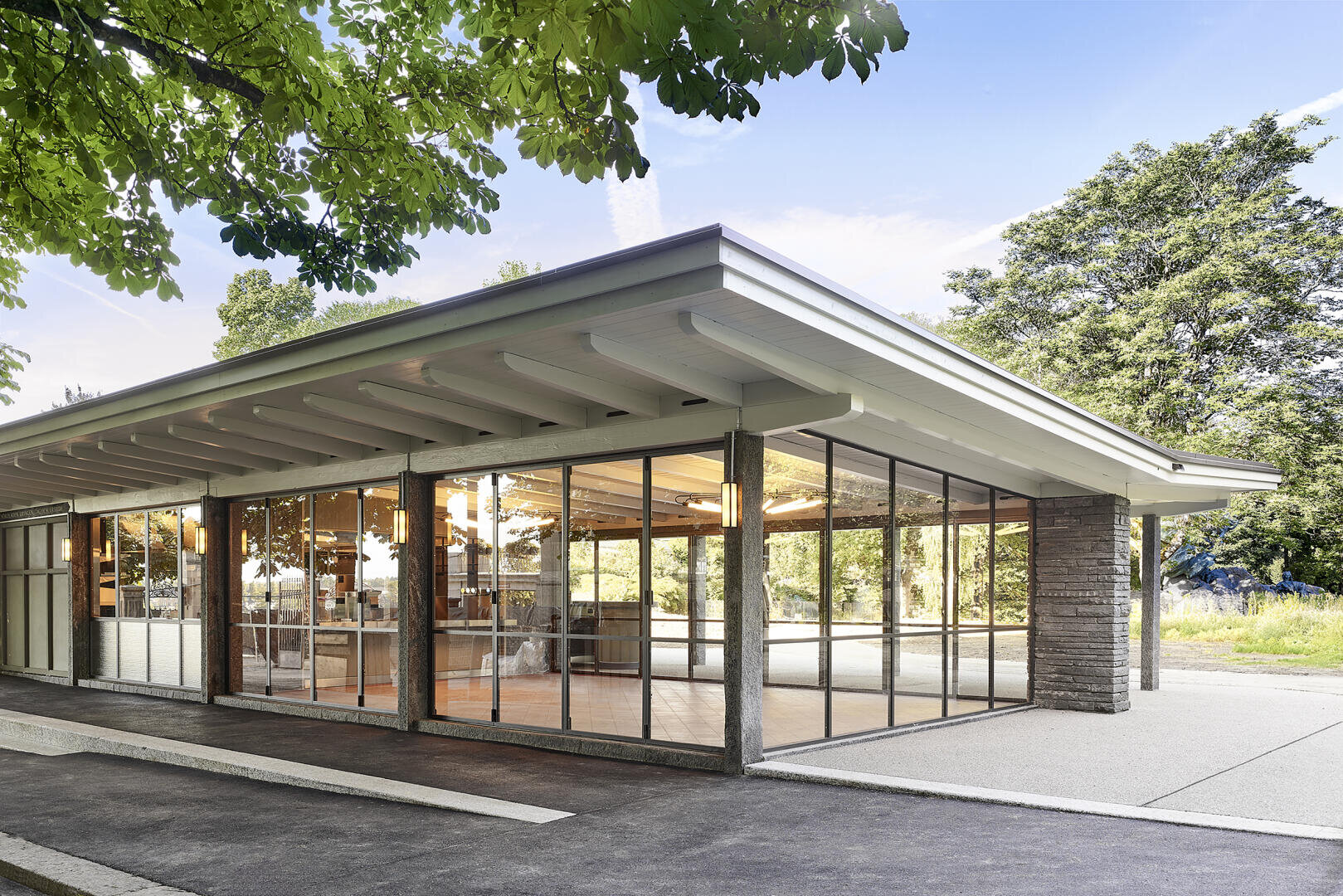 Ein modernes Gebäude mit großen Glaswänden und einem flachen, überhängenden Dach liegt inmitten von Bäumen und gibt den Blick frei auf das helle und geräumige Innere. Beton und natürliche Materialien vervollständigen das schlichte, offene Design.