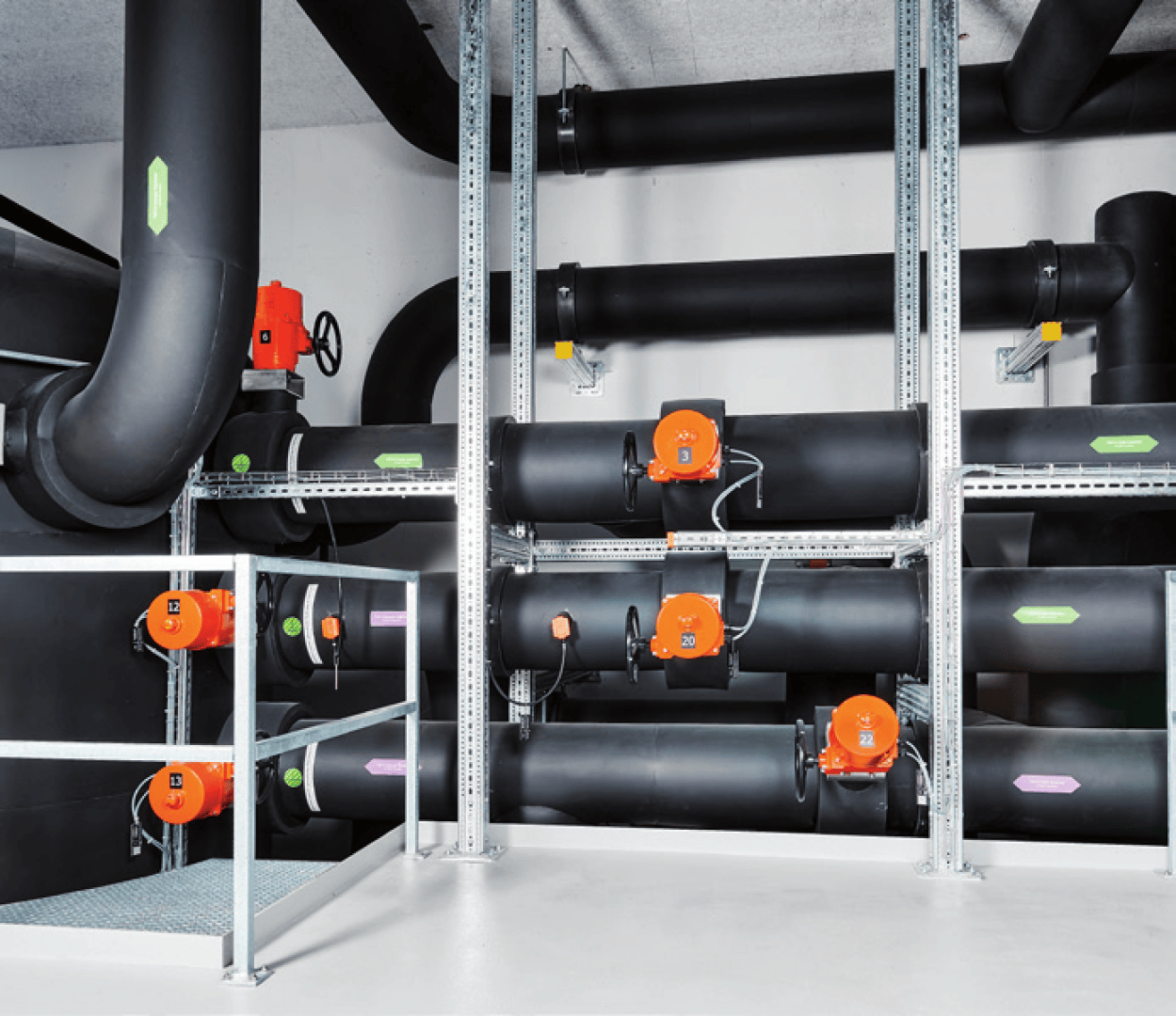 Une pièce propre et industrielle avec de gros tuyaux noirs isolés et des actionneurs de vannes orange vif montés sur des cadres métalliques qui courent horizontalement et verticalement le long des murs et du plafond blancs.