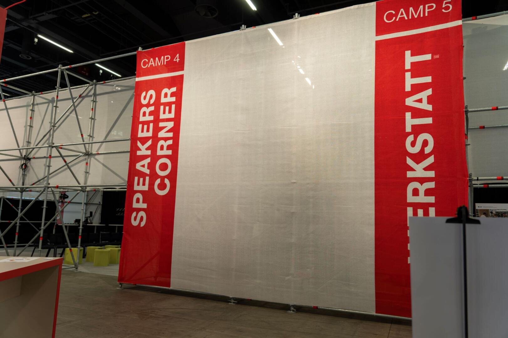 Eine große rot-weiße Trennwand an einem Veranstaltungsort zeigt die Worte SPEAKERS CORNER und WERKSTATT mit der Aufschrift CAMP 4 und CAMP 5 am oberen Rand, neben einem Metallgerüst.