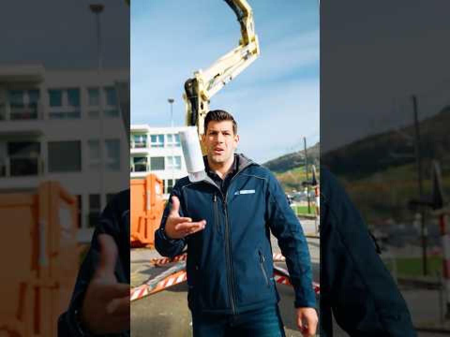 Ein Mann in einer blauen Jacke steht im Freien in der Nähe von Baumaschinen und Gebäuden und gestikuliert mit einer Hand. Im Hintergrund ist ein großer gelber Baggerarm zu sehen.