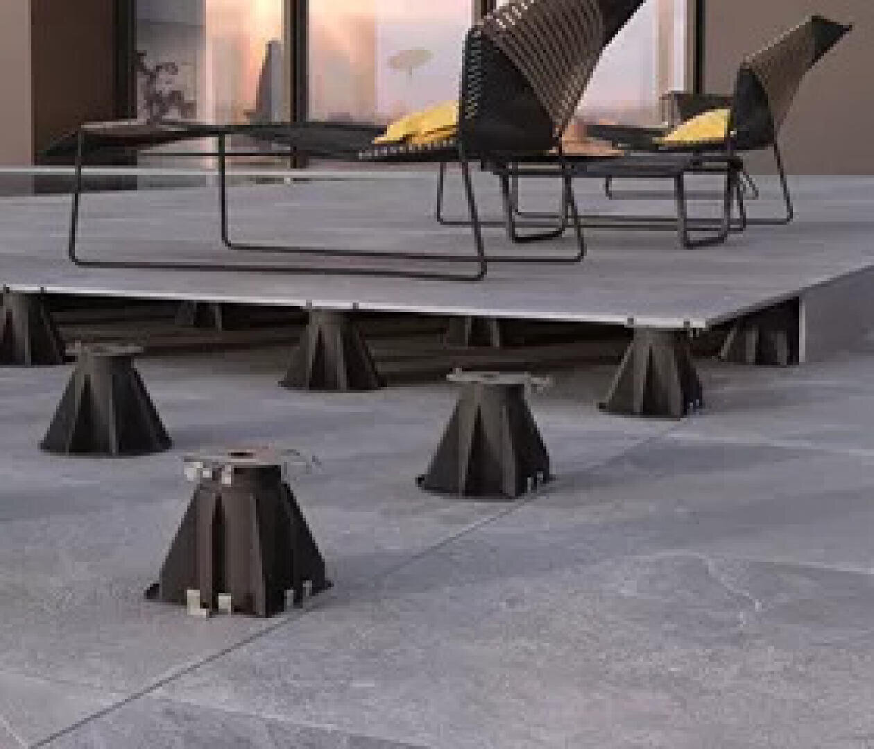 Un patio extérieur surélevé avec des dalles grises soutenues par des socles ajustables noirs, avec des chaises modernes noires et des coussins jaunes sur la surface surélevée.