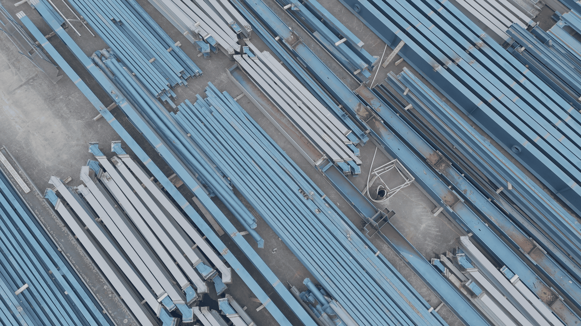Luftaufnahme von fein säuberlich gestapelten blauen und weißen Stahlträgern, die parallel auf einer Betonfläche angeordnet sind, möglicherweise auf einer Baustelle oder einem Lagerplatz.