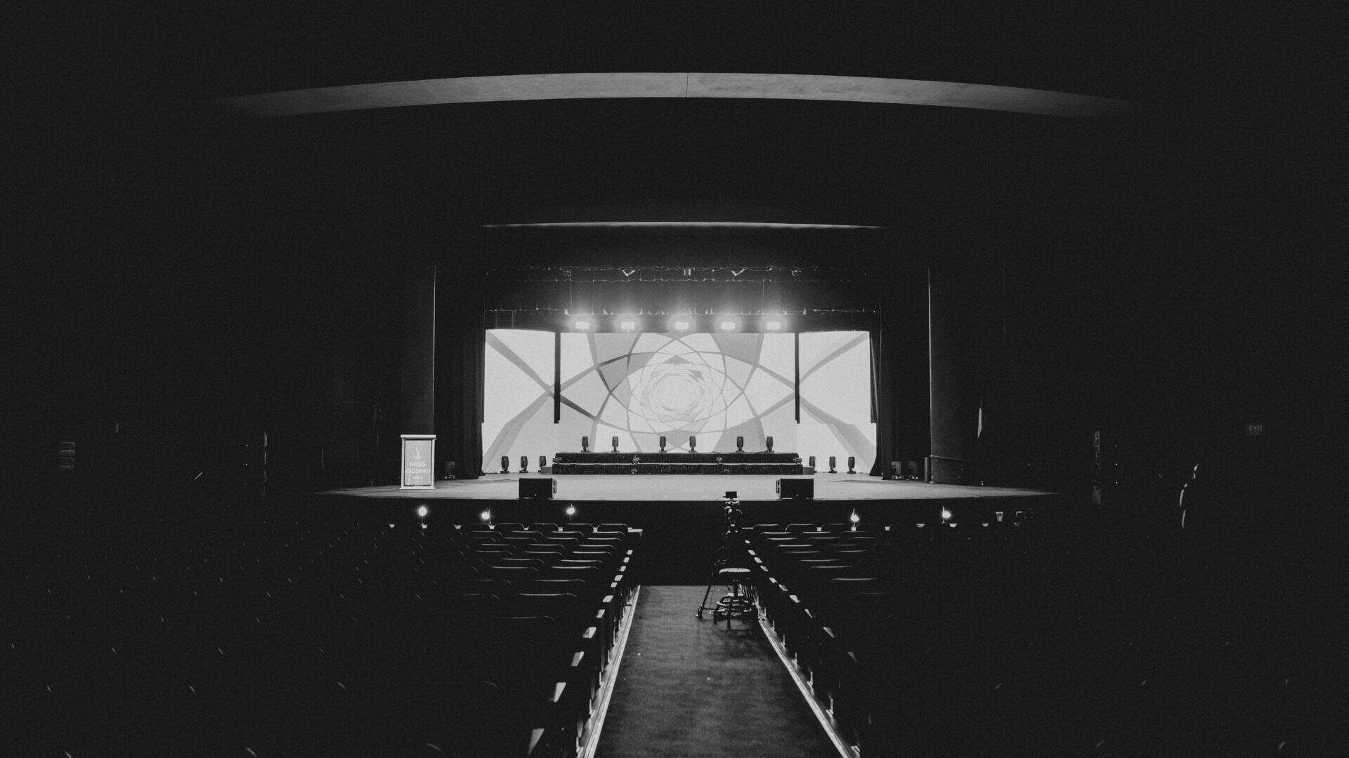 Ein Schwarz-Weiß-Foto eines leeren Theaters mit Sitzreihen vor einer hell erleuchteten Bühne, auf der eine große Leinwand mit abstrakten Mustern und ein langer Tisch mit Stühlen aufgebaut sind.