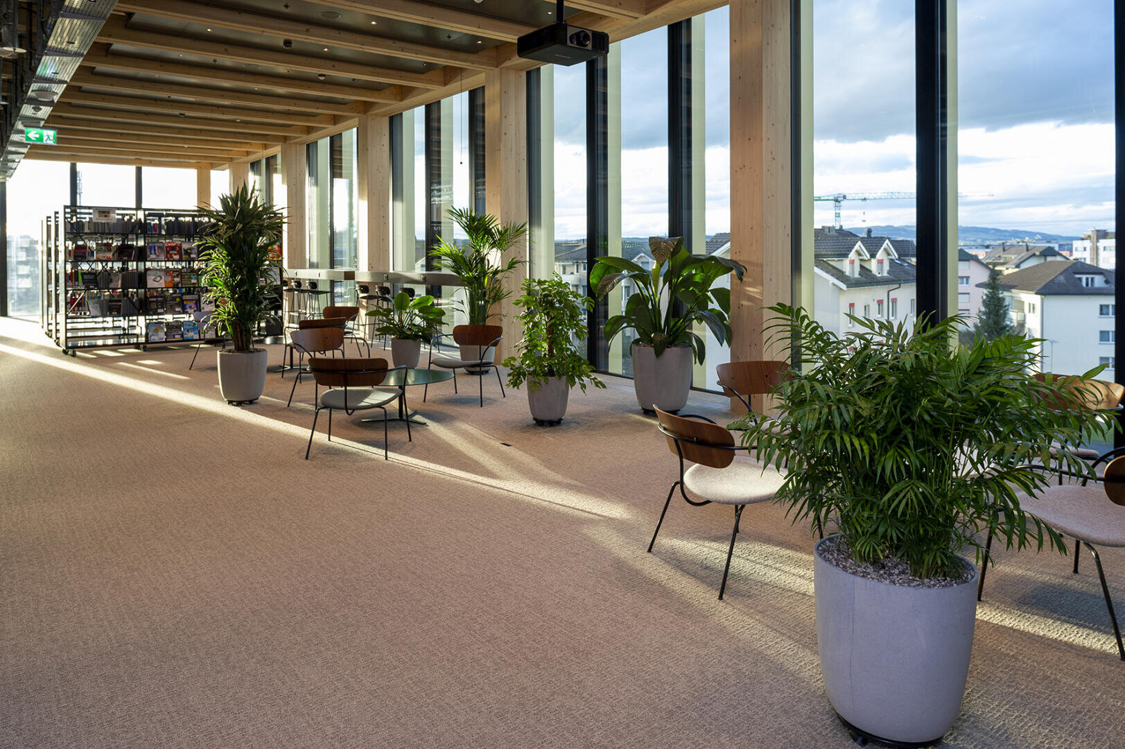 Un espace lounge moderne avec de grandes fenêtres, des poutres en bois, plusieurs petites tables et chaises et de nombreuses plantes vertes en pot. En arrière-plan, on peut voir des étagères avec des snacks et des boissons. Dehors, on peut voir les bâtiments de la ville.