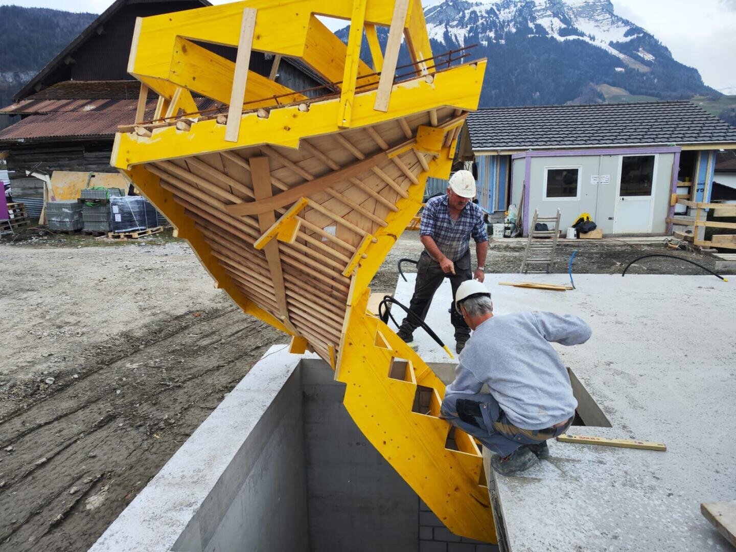 Zwei Bauarbeiter montieren auf einer Baustelle eine große, gelbe Holztreppe über einem Betonkeller, mit Bergen und traditionellen Gebäuden im Hintergrund.