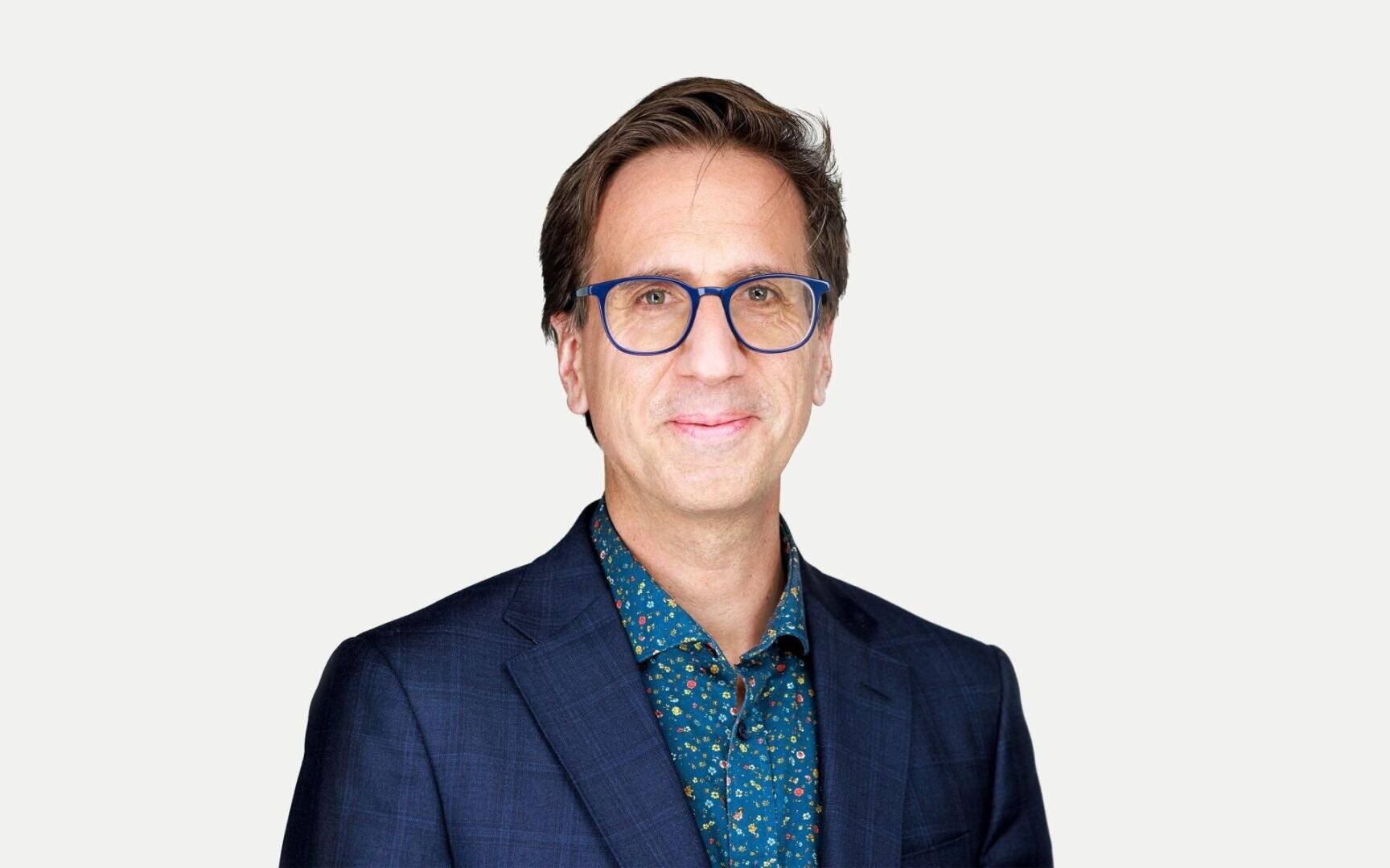 Ein Mann mit einer blauen Brille, einem marineblauen Blazer und einem blau gemusterten Hemd lächelt vor einem schlichten weißen Hintergrund in die Kamera.