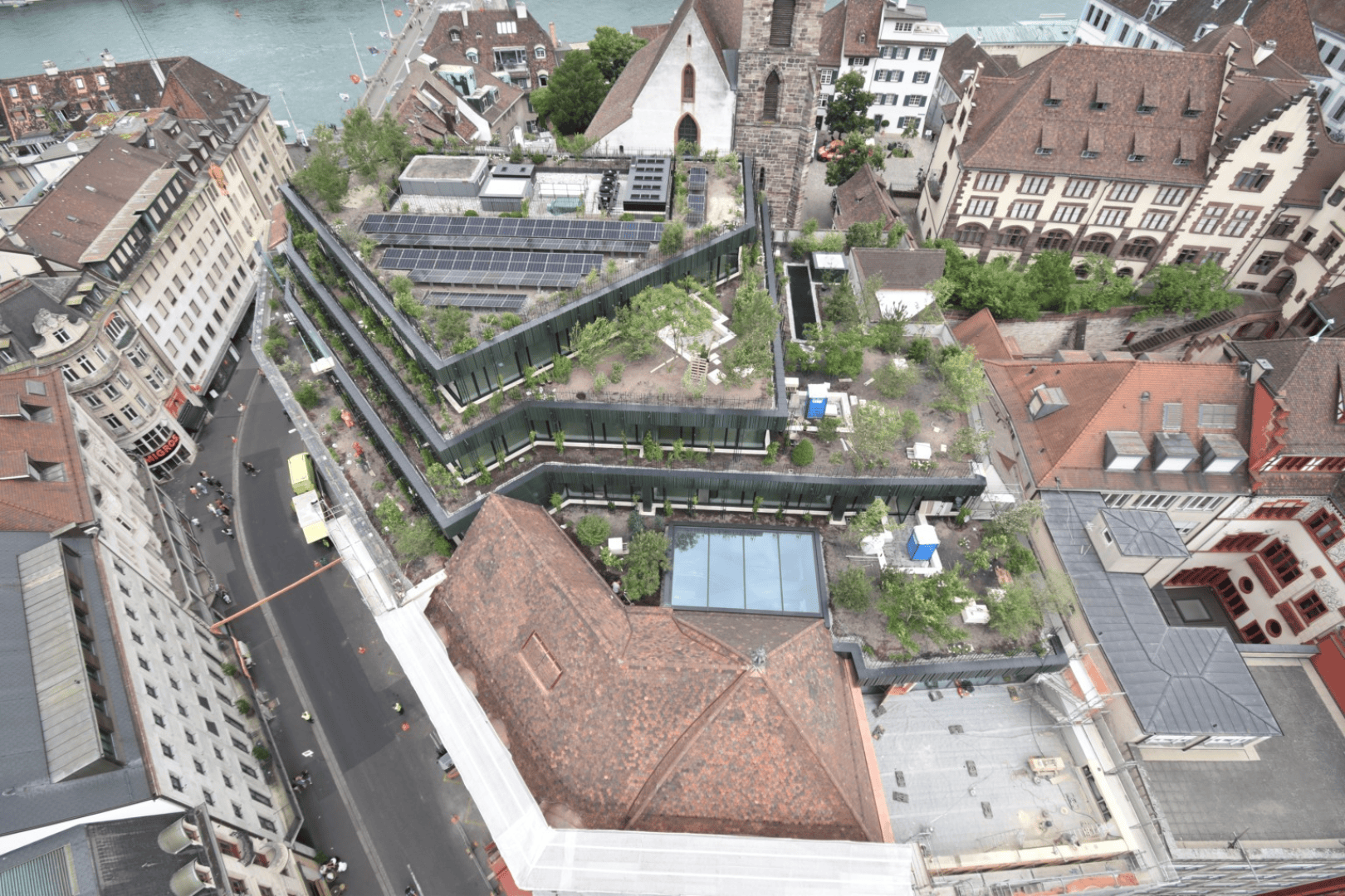 Luftaufnahme eines Stadtgebiets mit einem mehrstöckigen Gebäude mit Dachgärten und Sonnenkollektoren, umgeben von historischen Gebäuden und einem Fluss im Hintergrund.