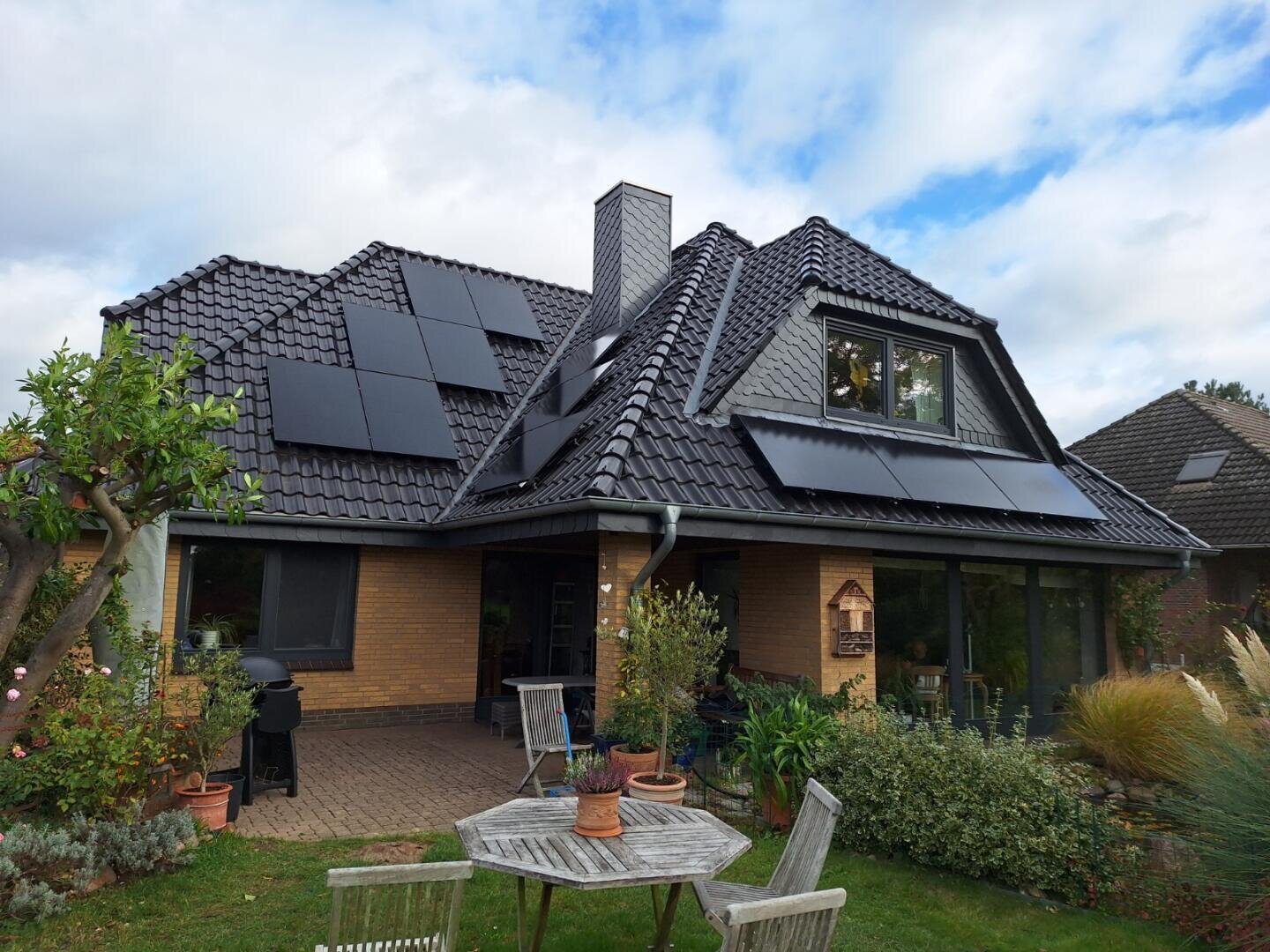 Ein modernes Backsteinhaus mit schwarzen Dachziegeln und mehreren auf dem Dach installierten Solarzellen, umgeben von einem Garten mit Gartenmöbeln, Pflanzen und Bäumen unter einem teilweise bewölkten Himmel.