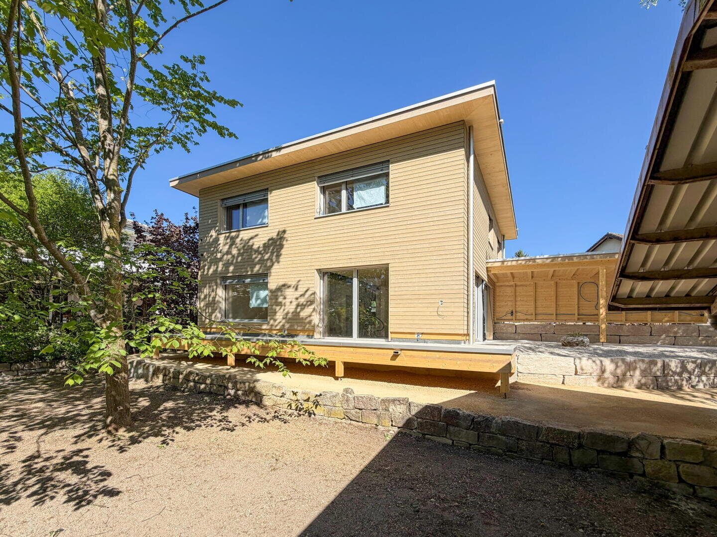 Ein modernes zweistöckiges Holzhaus mit großen Fenstern, einem Flachdach und einer erhöhten Holzterrasse, umgeben von Bäumen und einer Steinmauer unter einem klaren blauen Himmel.