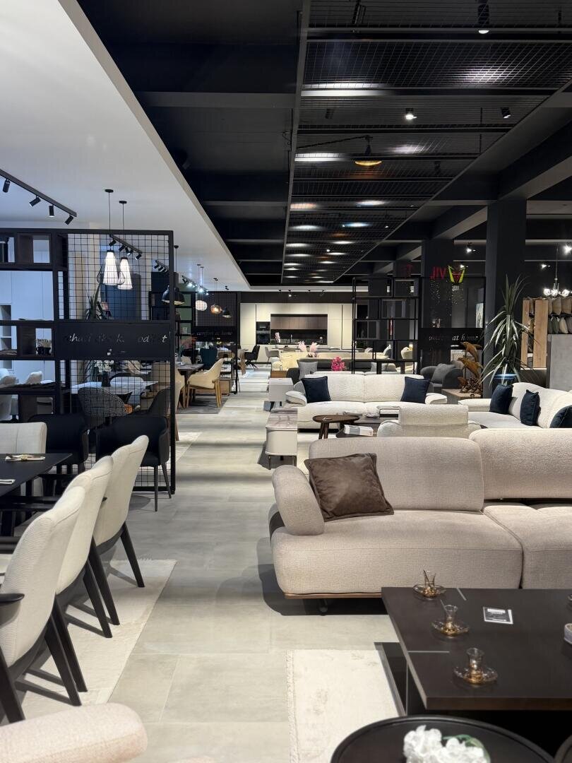 Ein moderner Möbelschauraum mit beigen Sofas, Sesseln und dunklen Holztischen, die ordentlich angeordnet sind. Der Raum ist mit Oberlichtern gut beleuchtet, und im ganzen Geschäft gibt es dekorative Accessoires und Pflanzen.