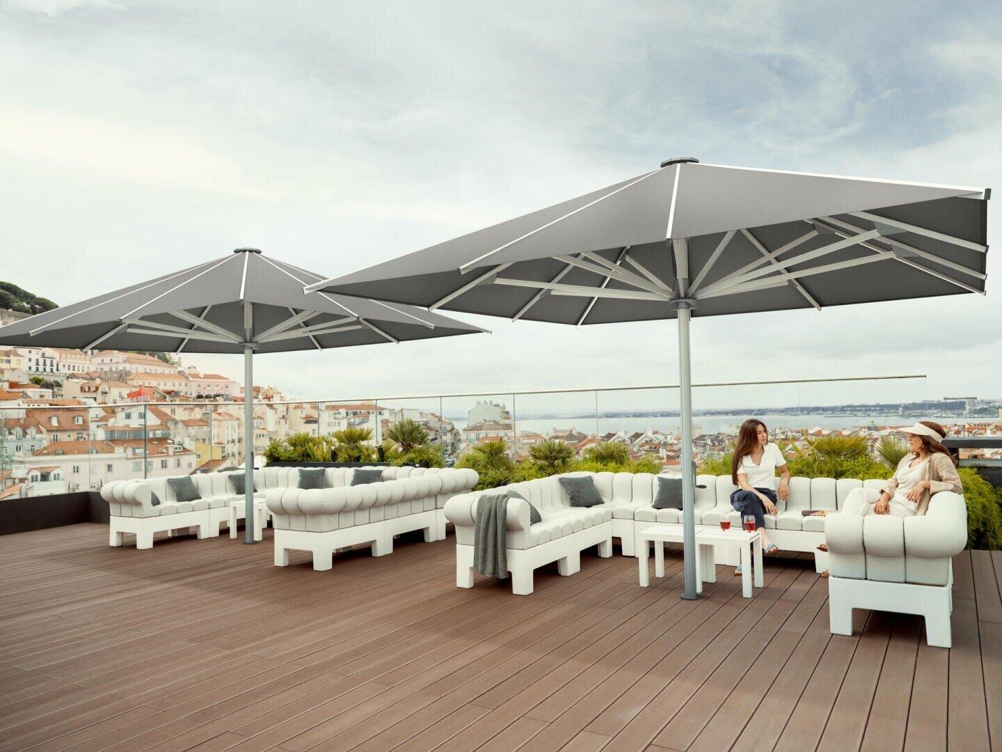 Zwei Frauen sitzen auf modernen weißen Sofas unter großen grauen Sonnenschirmen auf einer geräumigen Holzterrasse und blicken auf eine malerische Stadtlandschaft mit Gebäuden in den Bergen und Grünflächen im Hintergrund.