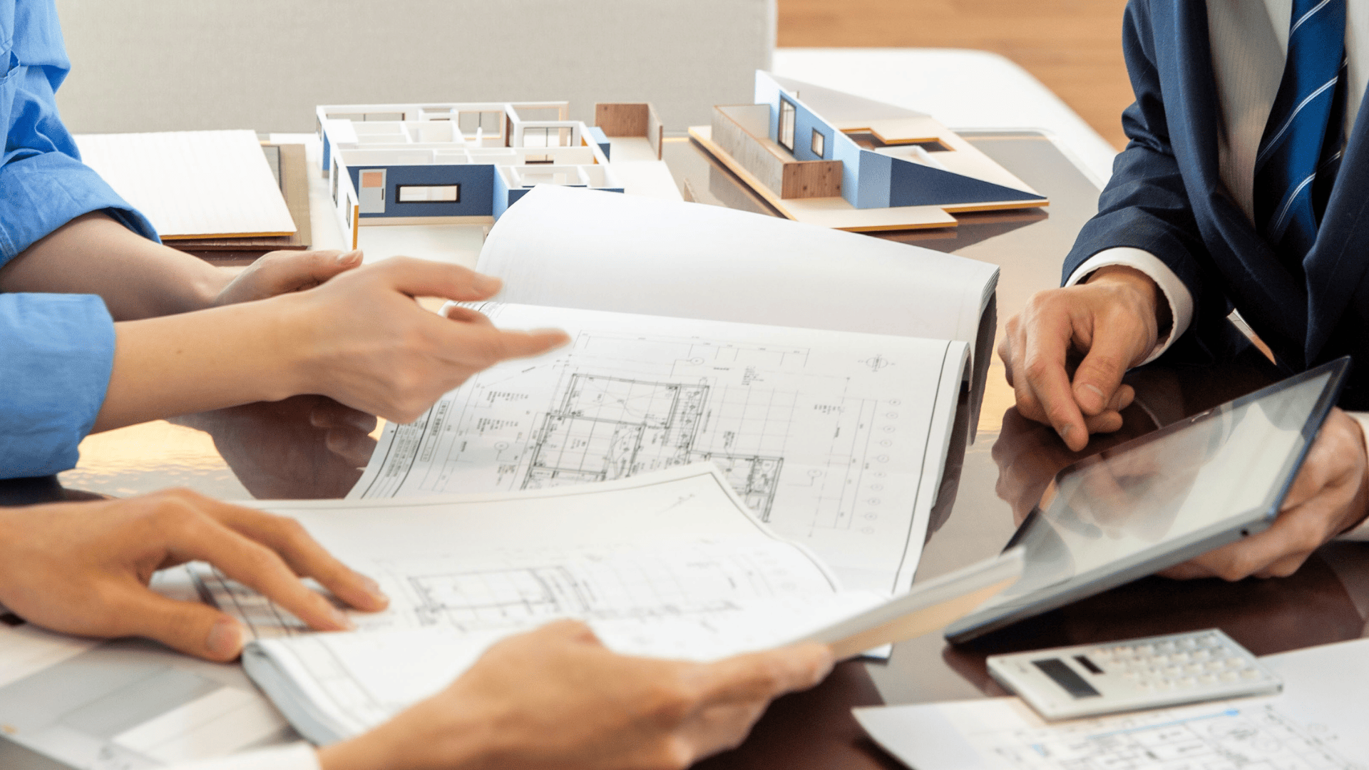 Drei Personen, die an einem Tisch mit architektonischen Modellen, einem Taschenrechner und einem Tablet architektonische Entwürfe und Dokumente prüfen, was auf eine Besprechung oder Diskussion über die Gestaltung oder Planung von Gebäuden hindeutet.