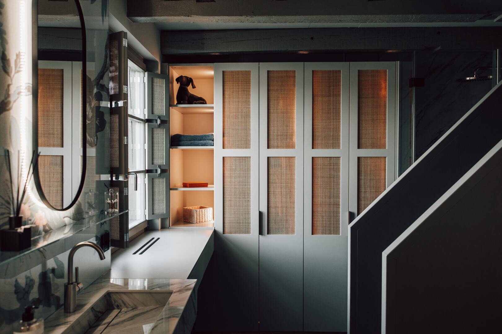 Modernes Badezimmer mit Marmorarbeitsplatte, Waschbecken und Spiegel auf der linken Seite, eingebauten Regalen mit Handtüchern und einem Korb am Fenster und hohen Schränken mit gewebter Verkleidung, die teilweise von einer weichen, warmen Beleuchtung beleuchtet werden.