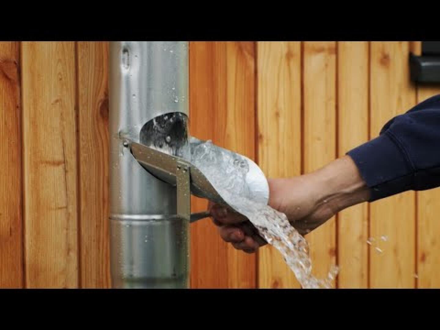 Eine Person fängt mit einer Metallschaufel das Wasser auf, das aus einem an einer Holzwand befestigten Fallrohr aus verzinktem Stahl fließt.
