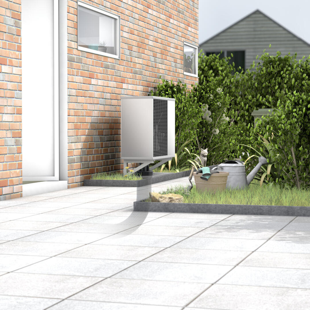 Eine Wärmepumpeneinheit, die außerhalb eines Backsteinhauses auf einer Terrasse montiert ist, mit einer Gießkanne, Gartengeräten und Pflanzen in der Nähe. Im Hintergrund sind ein Wohngebäude und Büsche zu sehen.
