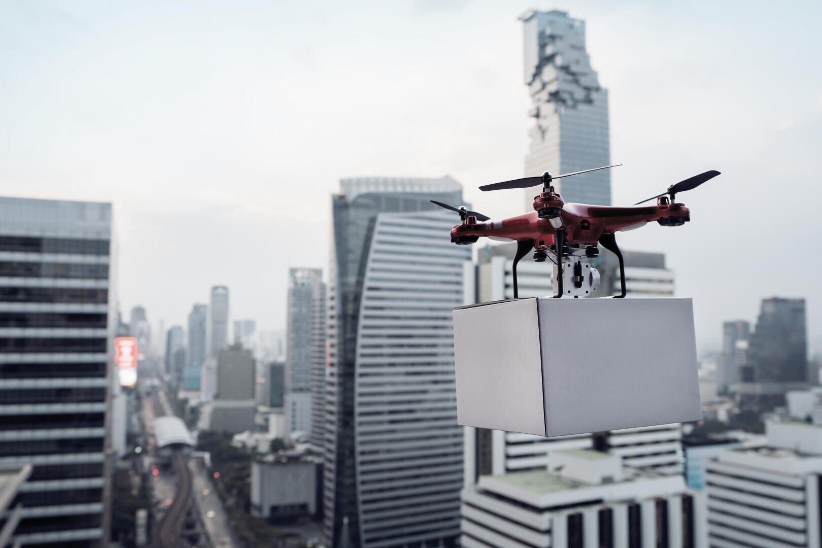 Eine rote Drohne, die ein rechteckiges Paket trägt, fliegt über eine Stadtlandschaft mit hohen modernen Gebäuden und einem bewölkten Himmel im Hintergrund.