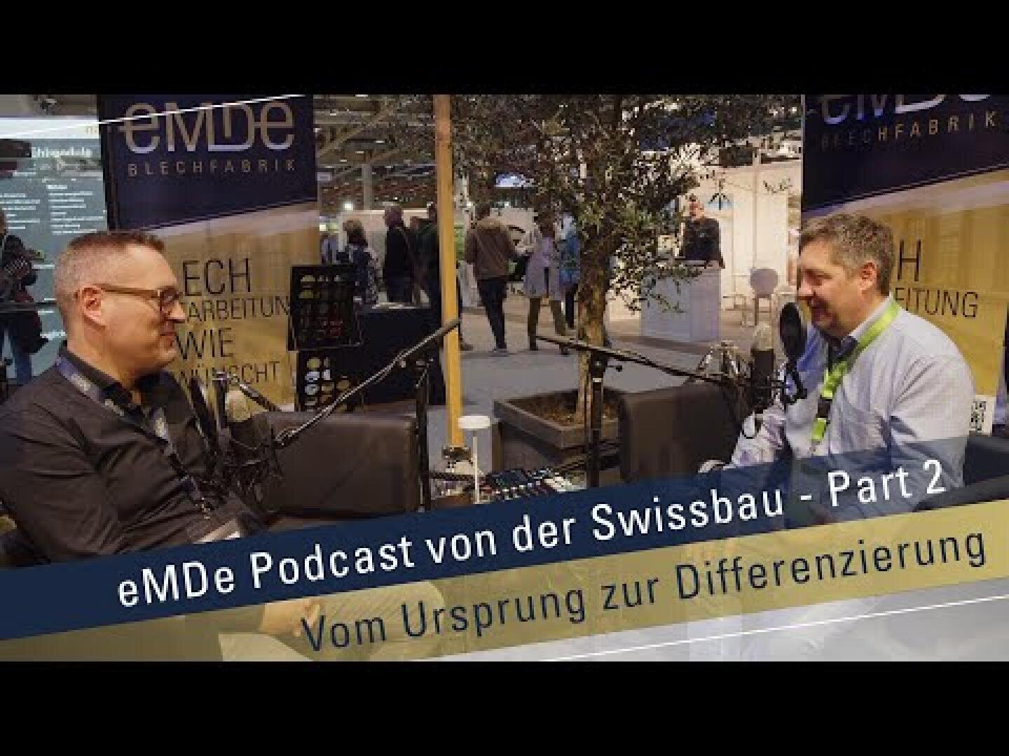 Zwei Männer sitzen an einem Tisch mit Mikrofonen und nehmen auf einer Messe einen Podcast auf. Der Text lautet: eMDe Podcast von der Swissbau - Teil 2 Vom Ursprung zur Differenzierung. Im Hintergrund ist eine belebte Messehalle zu sehen.