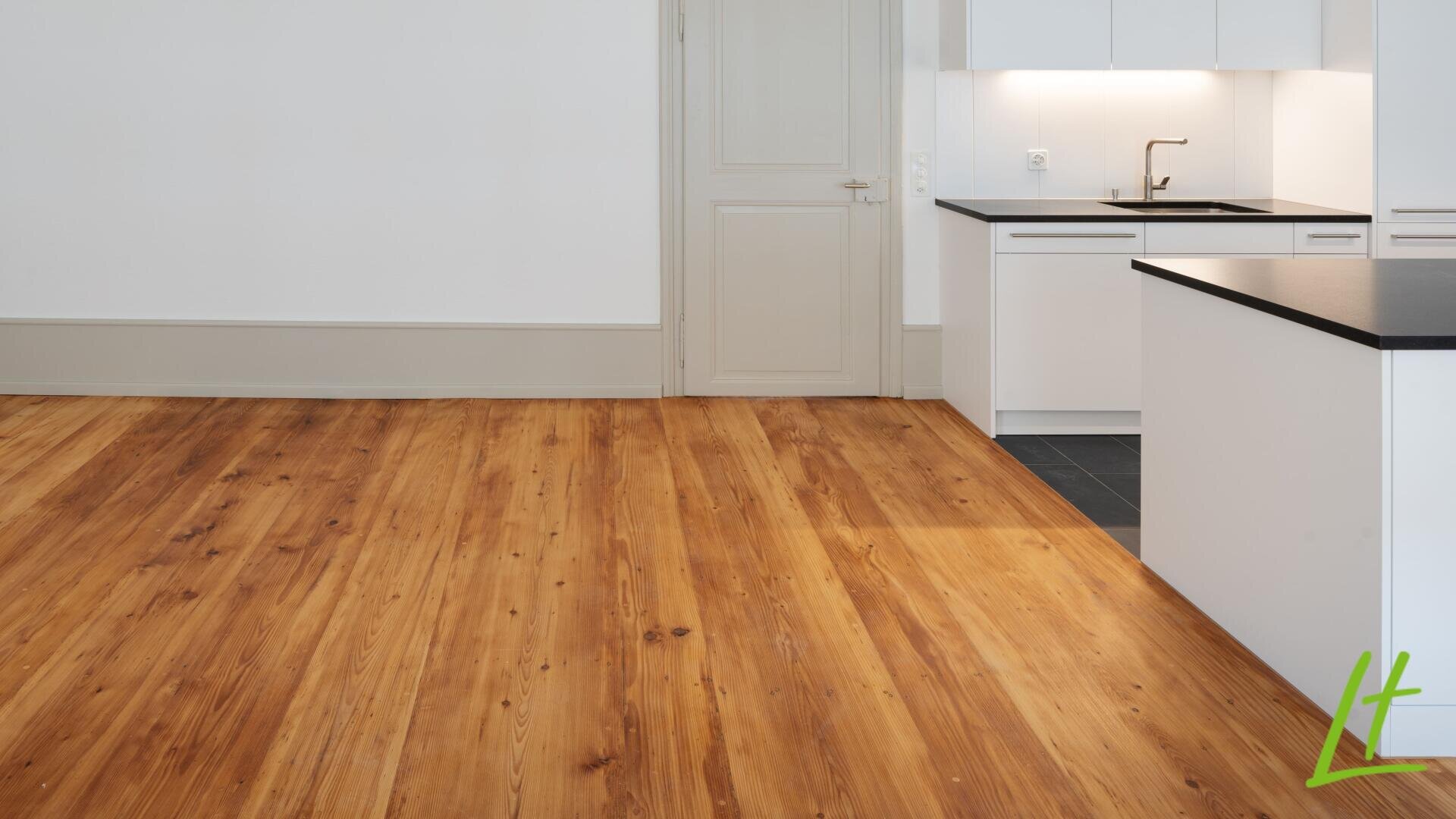 Eine moderne Küche mit weißen Schränken, einer schwarzen Arbeitsplatte, einer Spüle und einem Holzboden; der Raum hat eine geschlossene Tür und ein Logo mit grünen und gelben Linien in der unteren rechten Ecke.