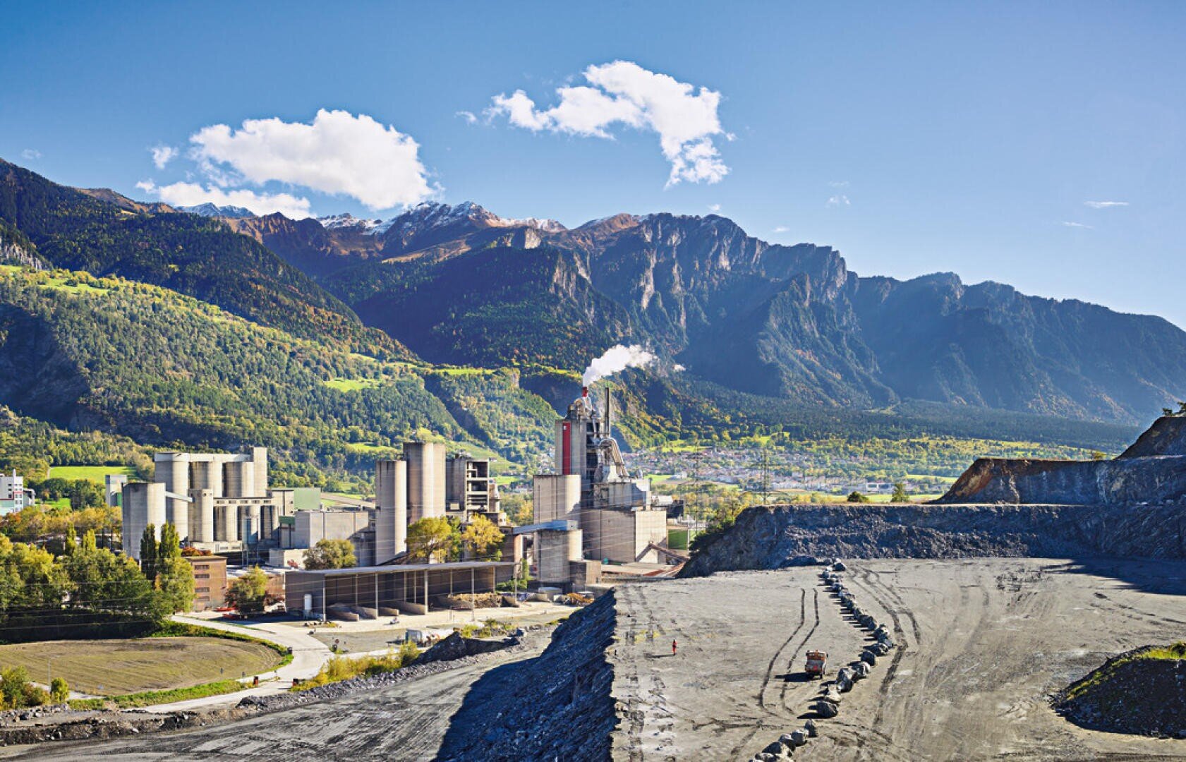 Eine Zementfabrik raucht inmitten grüner Hügel und Berge unter einem blauen, wolkenverhangenen Himmel; ein felsiger, karger Vordergrund kontrastiert mit der üppigen Landschaft und den fernen Schneegipfeln.