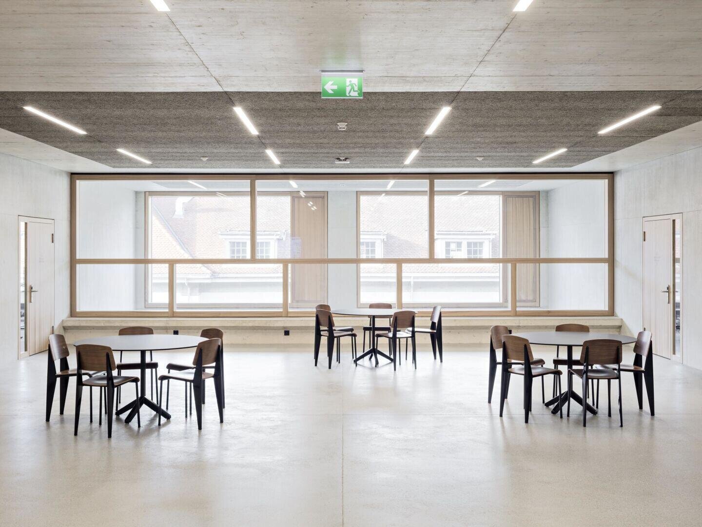 Ein geräumiger, moderner Raum mit vier Tischen und mehreren Stühlen, großen Fenstern, die natürliches Licht hereinlassen, hellen Wänden und Böden sowie einem Notausgangsschild an der Decke.