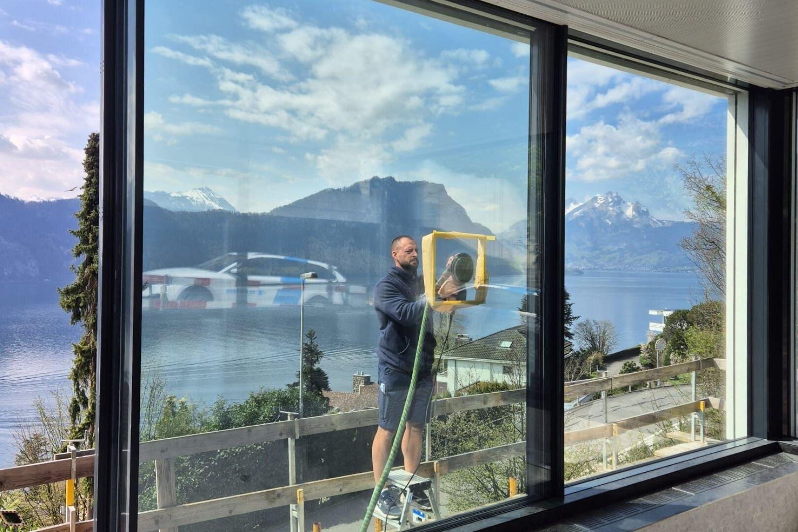 Un homme se tient dehors sur une échelle et nettoie de grandes fenêtres en verre avec une raclette. L'arrière-plan pittoresque montre des montagnes enneigées, un lac et un ciel partiellement nuageux.