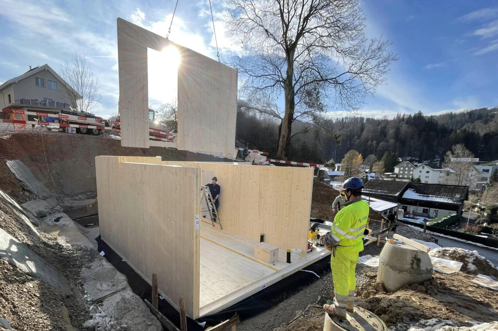 Un chantier sur lequel des ouvriers assemblent un bâtiment en bois ; une grue soulève un grand panneau mural en bois au-dessus de sa tête, tandis qu'un ouvrier vêtu de jaune vif observe. En arrière-plan, on aperçoit des maisons, des arbres et un ciel bleu.