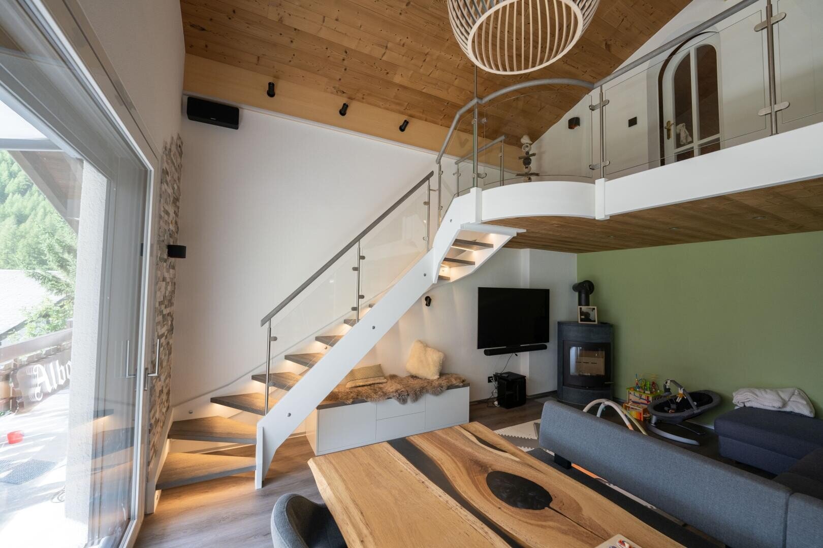 Salon moderne avec plafond en bois, escalier en verre, table à manger en bois clair, TV, cheminée, jouets pour enfants et vue sur un balcon. La lumière naturelle remplit la pièce et souligne l'aménagement moderne.