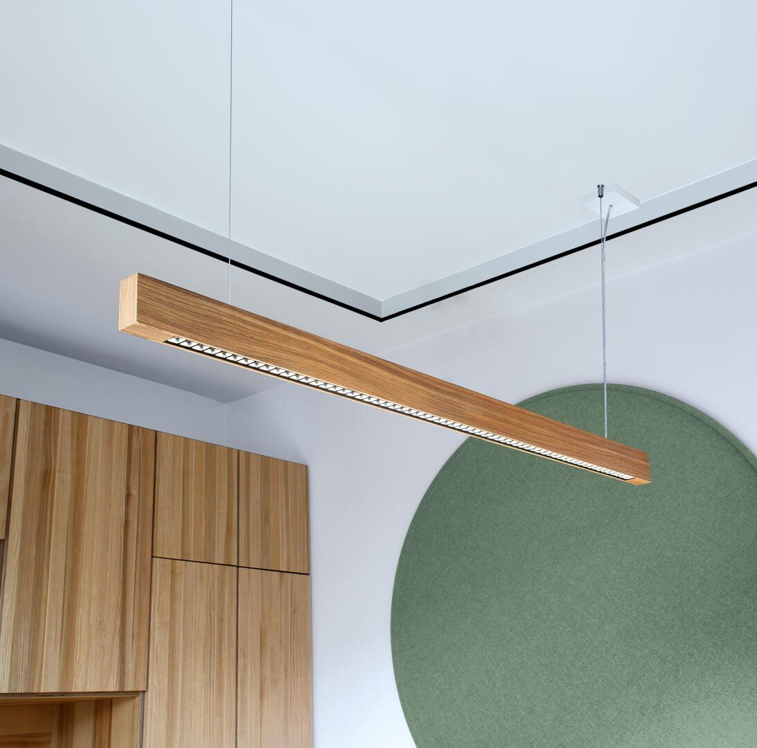 Eine moderne, geradlinige Pendelleuchte aus Holz hängt an zwei dünnen Kabeln von der Decke über einem Raum mit hellen Holzschränken und einem großen grünen runden Wandelement.