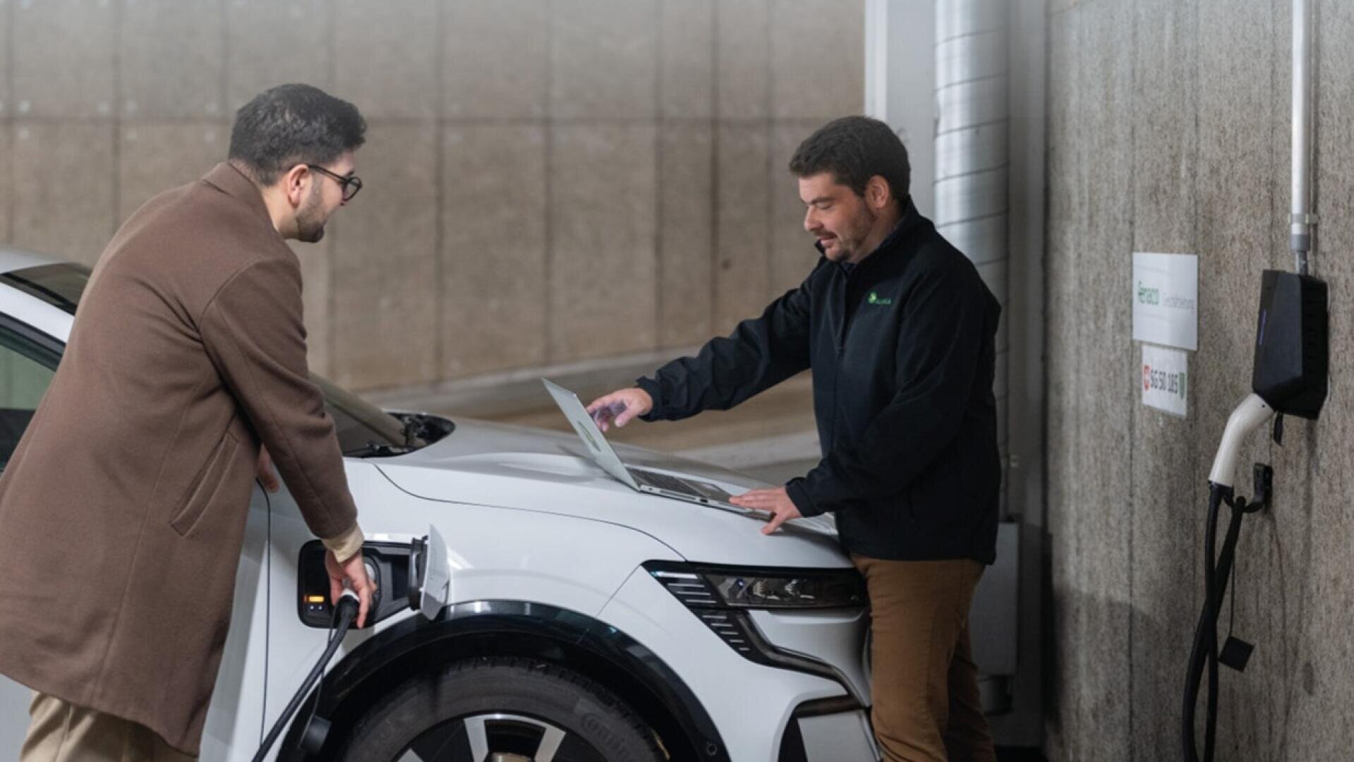Zwei Männer stehen neben einem weißen Elektroauto an einer Ladestation in einem Innenraum; ein Mann benutzt einen Laptop auf der Motorhaube des Autos, während der andere beobachtet, und ein Ladekabel ist an das Fahrzeug angeschlossen.