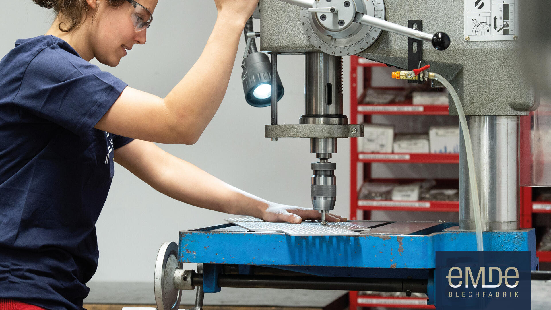 Eine Person arbeitet mit einer Bohrmaschine an einem Blech in einer Werkstatt. Im Hintergrund sind Schubladen mit organisierten Teilen zu sehen. Das Logo eMDe Blechfabrik erscheint in der unteren rechten Ecke.