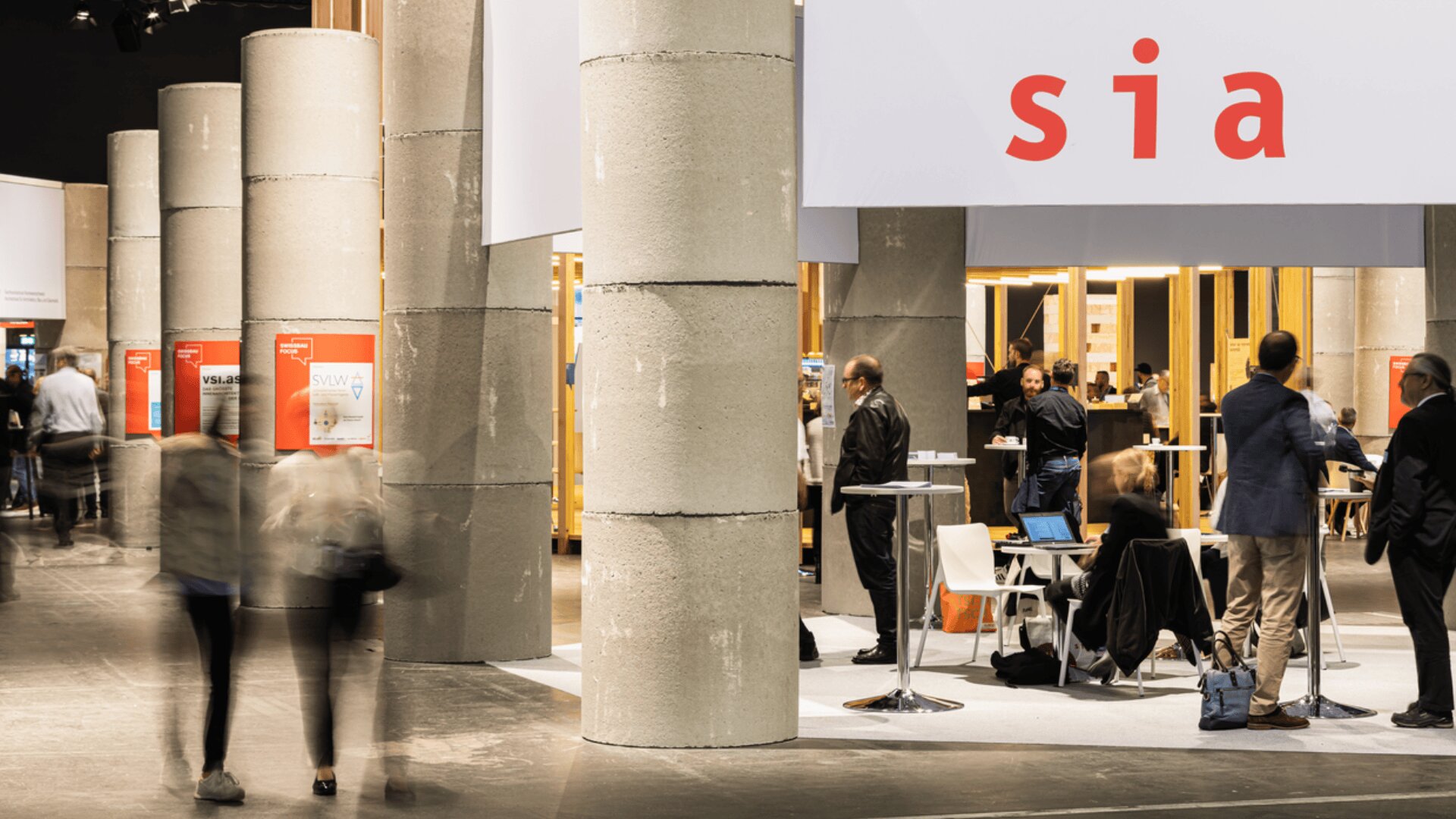 Des personnes se rassemblent et marchent autour d'un espace d'exposition intérieur moderne avec de grands piliers en béton et un stand présentant les lettres rouges sia sur une bannière blanche. Certaines personnes discutent tandis que d'autres se déplacent dans l'espace.
