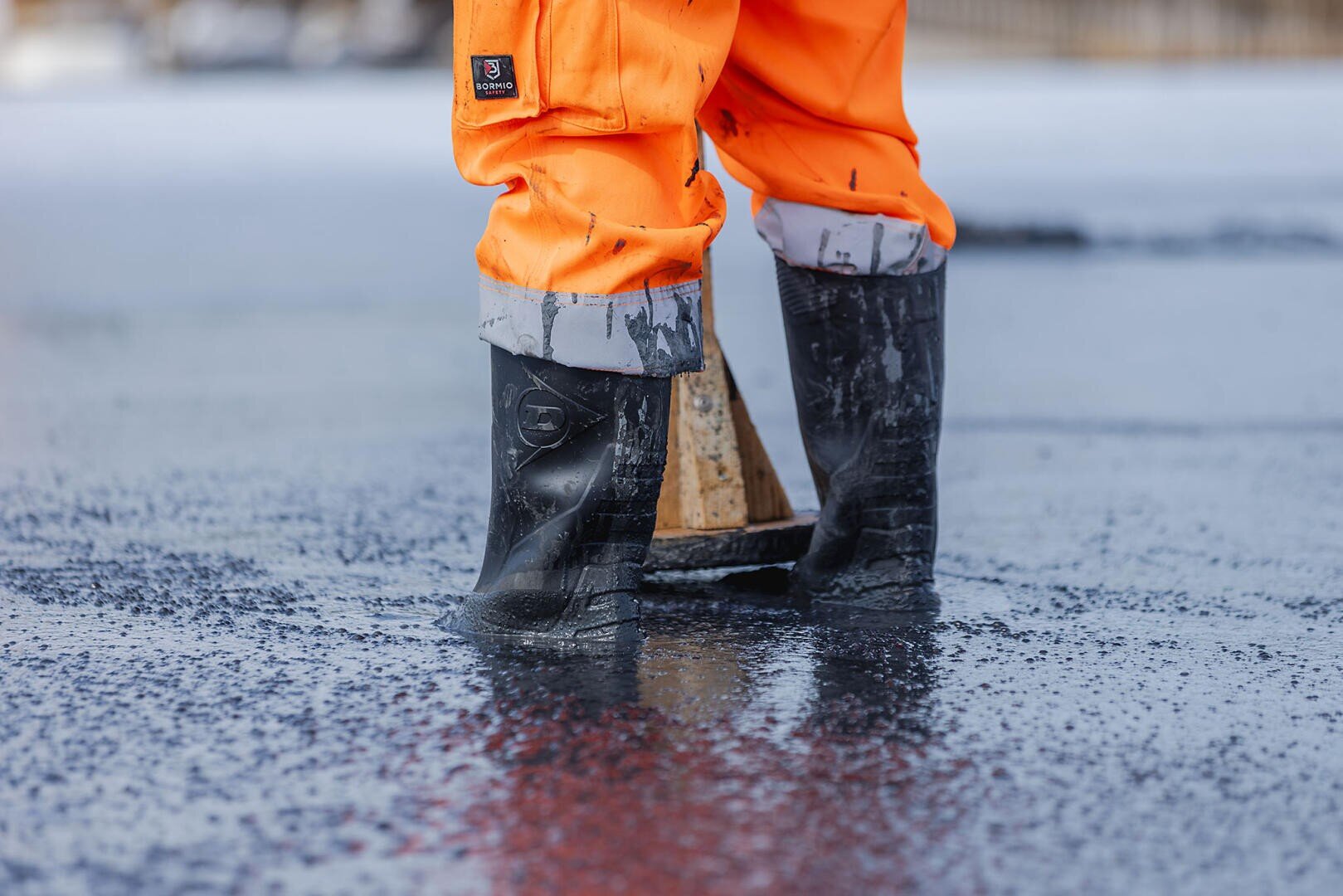 Eine Person, die eine orangefarbene wasserdichte Hose und schwarze Gummistiefel trägt, steht im flachen Wasser, wobei die Unterschenkel und Füße sichtbar sind. Die Wasseroberfläche spiegelt sich und erscheint kalt, möglicherweise eisig.