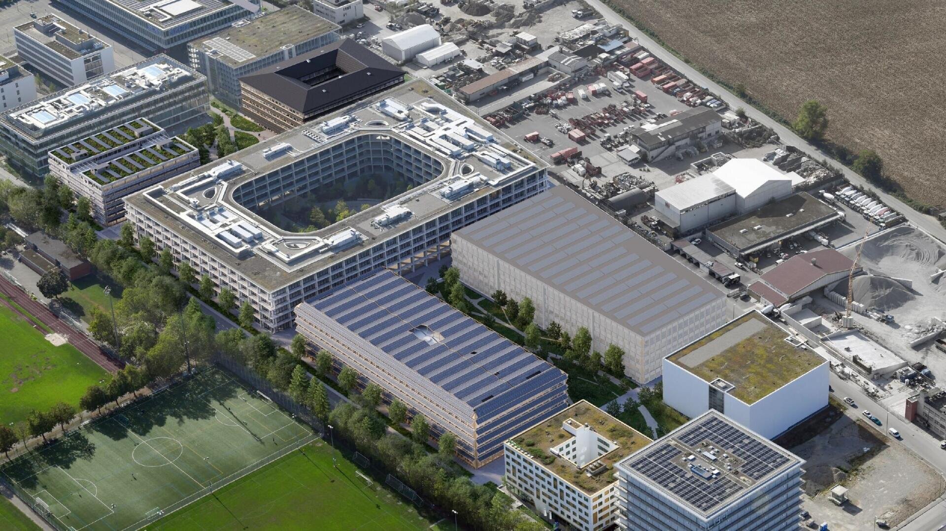 Luftaufnahme eines modernen Büro- und Industriekomplexes mit mehreren großen rechteckigen Gebäuden, Grünflächen, Bäumen, Parkplätzen und einem nahe gelegenen Sportplatz. Straßen und offene Flächen umgeben das Gelände.