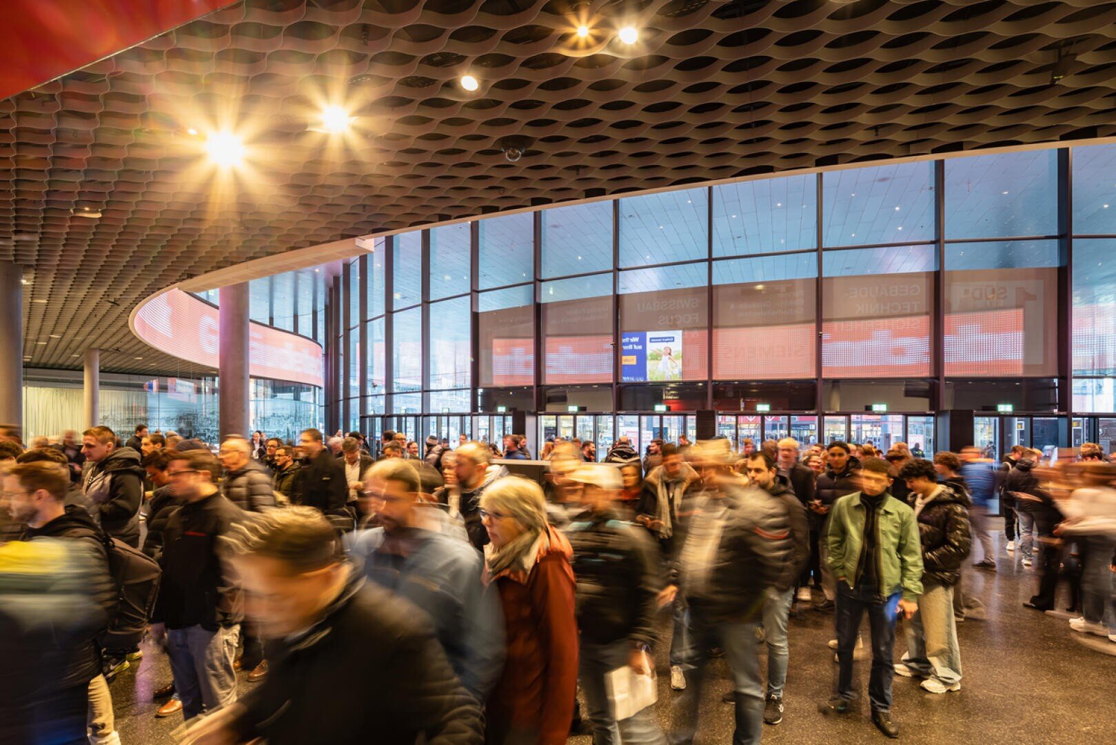 Eine große Menschenmenge, teilweise in Bewegungsunschärfe, geht in ein modernes Gebäude mit Glaswänden, hohen Decken und heller Beleuchtung. Die Szene wirkt geschäftig und energiegeladen, möglicherweise bei einem Kongress oder einer öffentlichen Veranstaltung.