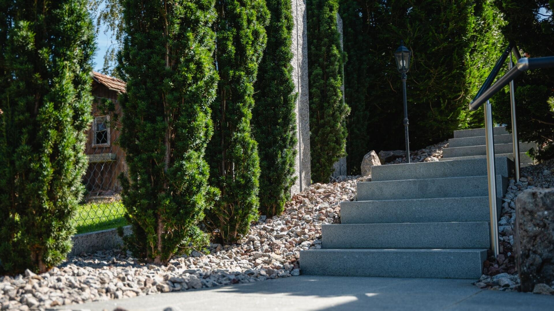 Eine Steintreppe im Freien, gesäumt von hohen grünen Büschen und dekorativen Steinen, führt neben einem Metallgeländer mit einer Gartenlaterne im Vintage-Stil in der Nähe nach oben. Das Sonnenlicht wirft Schatten auf den Weg.
