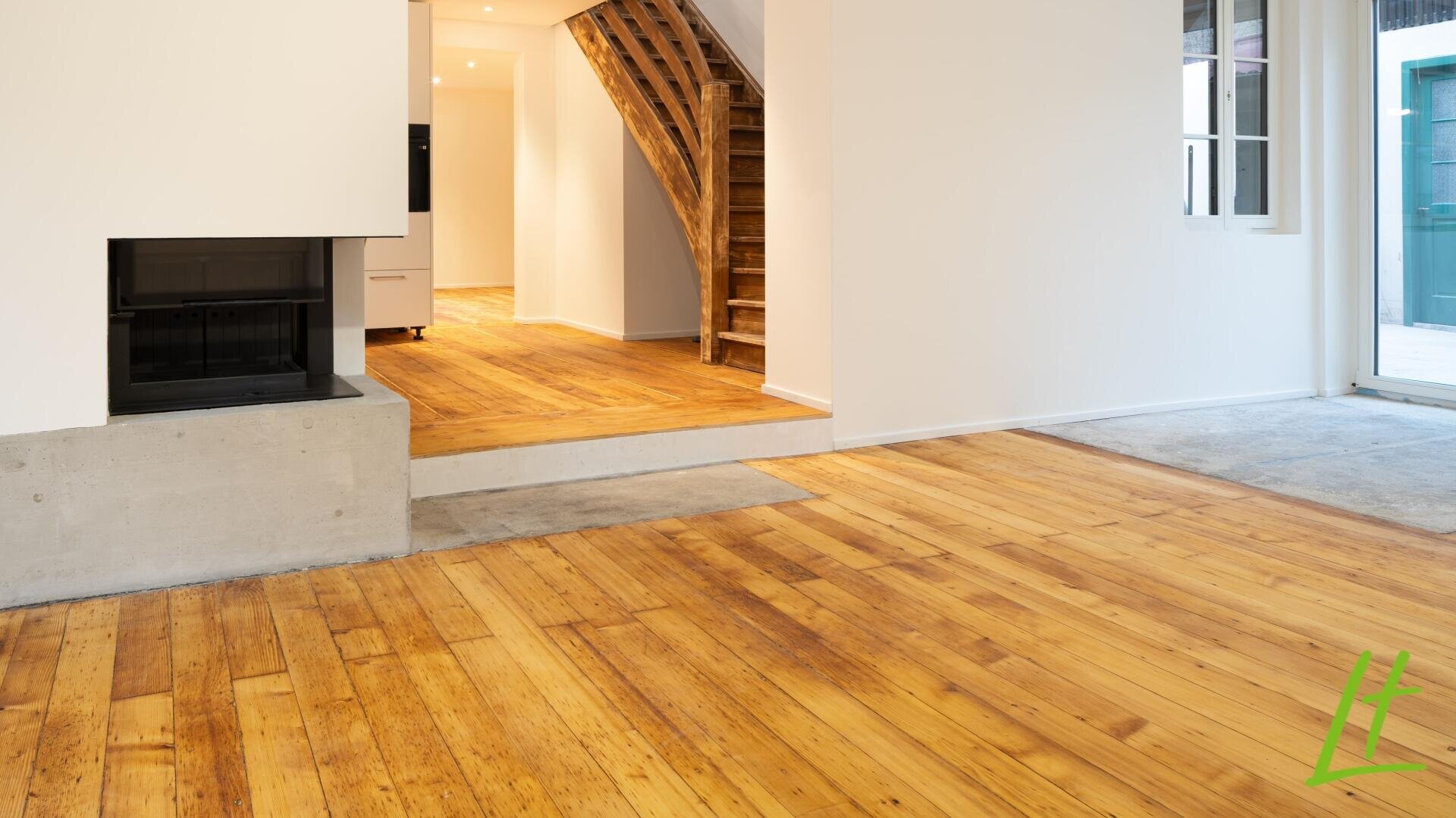 Helles Interieur mit Holzböden, einem Kamin auf der linken Seite, einer Holztreppe in der Mitte, weißen Wänden und großen Fenstern, die natürliches Licht hereinlassen. Eine Betontreppe trennt zwei Bereiche des Wohnraums.