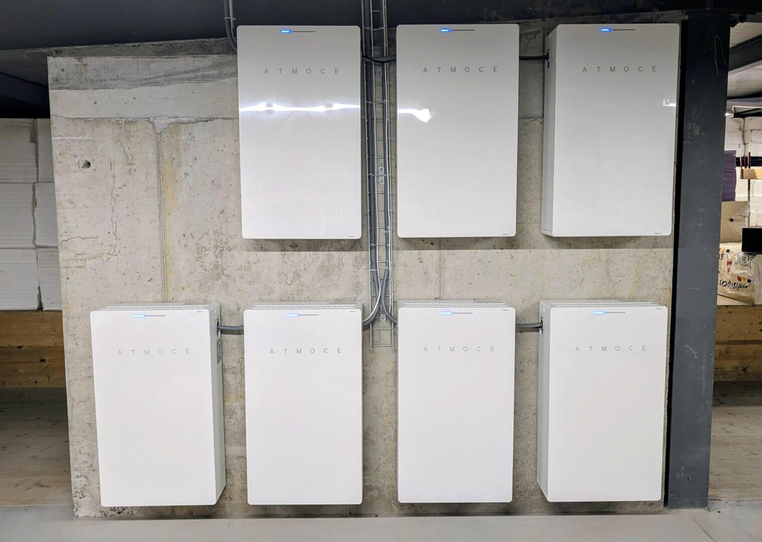 Sechs weiße, rechteckige Batteriesätze mit der Aufschrift "ATMOSCE" sind in zwei Dreierreihen an einer Betonwand angebracht und mit blauen Anzeigeleuchten versehen. Eine sichtbare Verkabelung verbindet die Einheiten miteinander. Der Raum hat unfertige Wände und Fußböden.